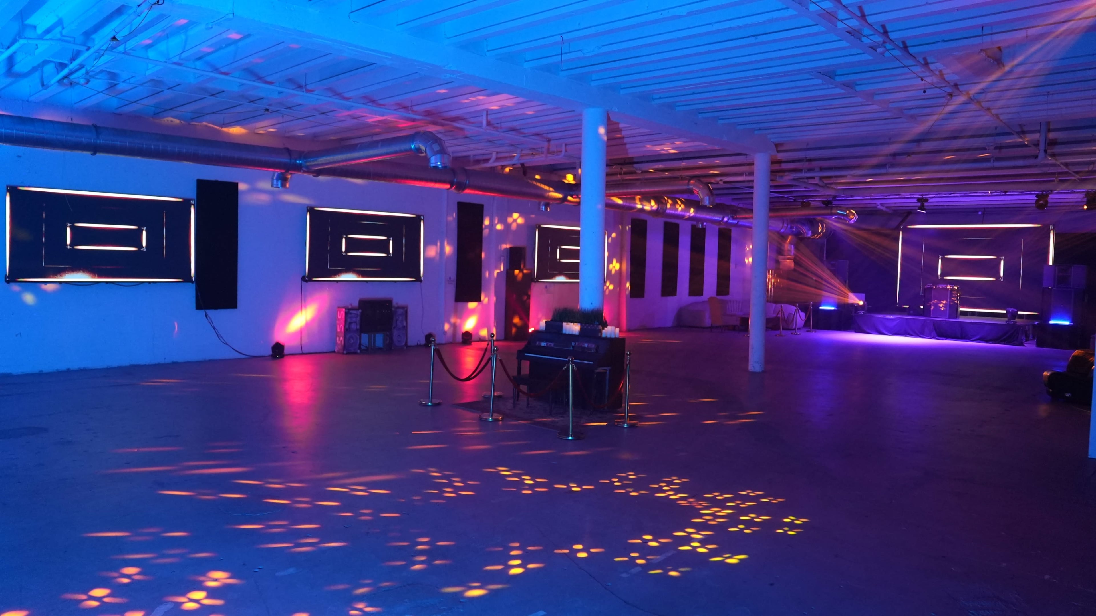The image shows a spacious, illuminated room with colorful lighting effects and multiple screens on the walls, indicating a setup for an event or performance.
