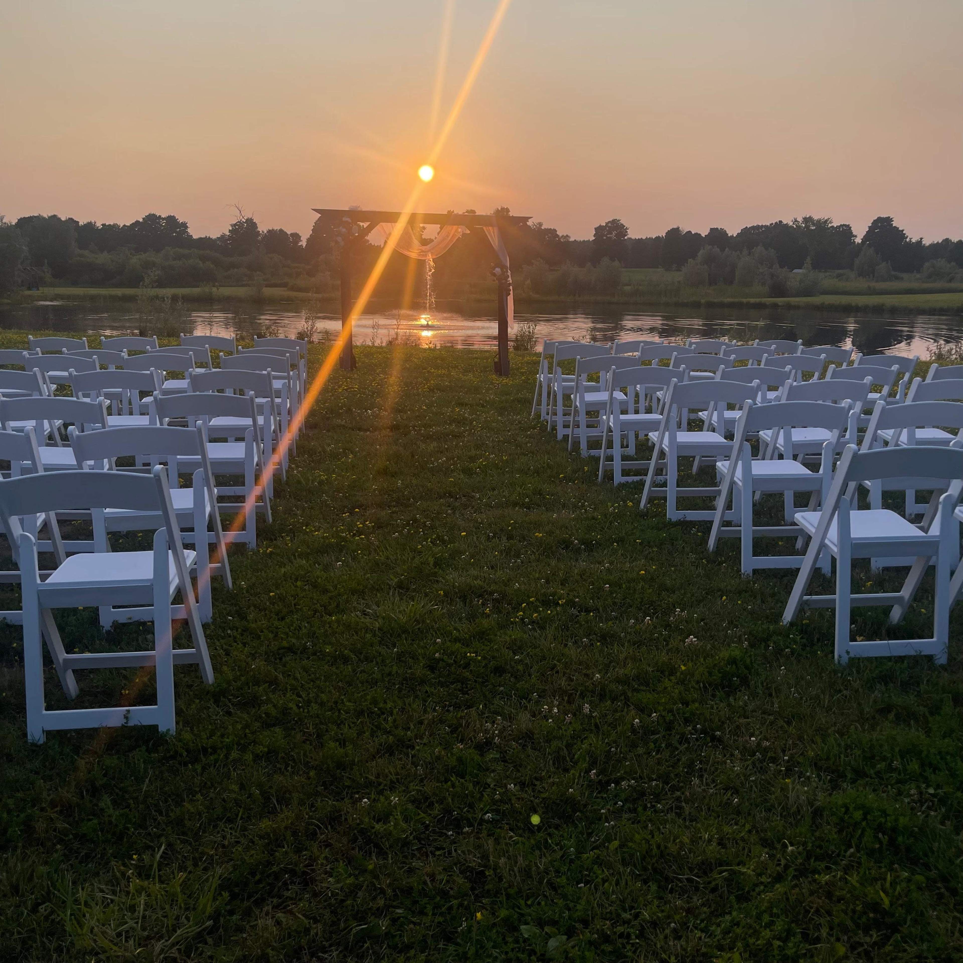 A row of white folding chairs is arranged on grass facing a wooden arch over a body of water at sunset.