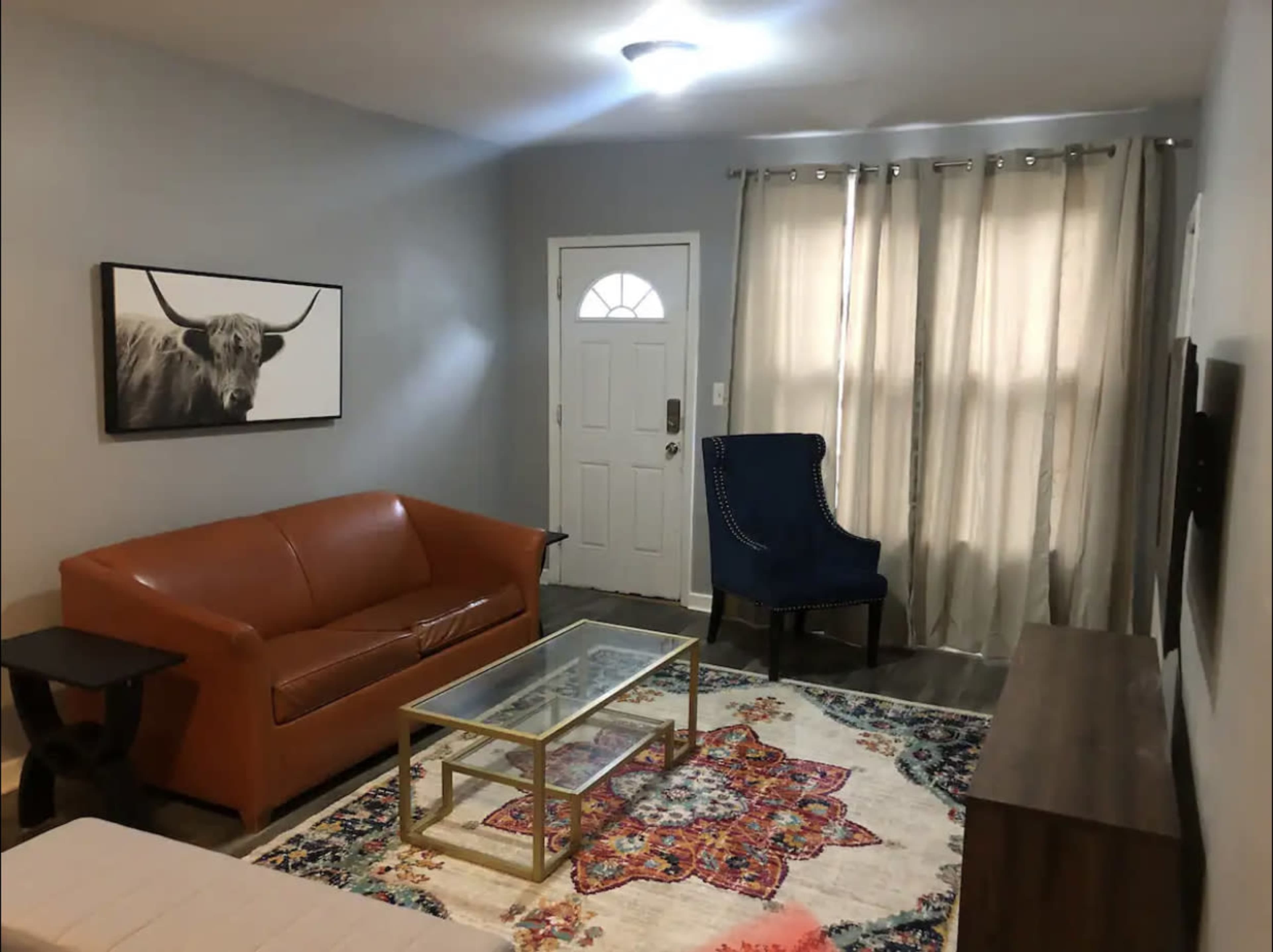 A living room with a brown leather sofa, a blue upholstered chair, a glass coffee table on a patterned rug, and a door with a window.
