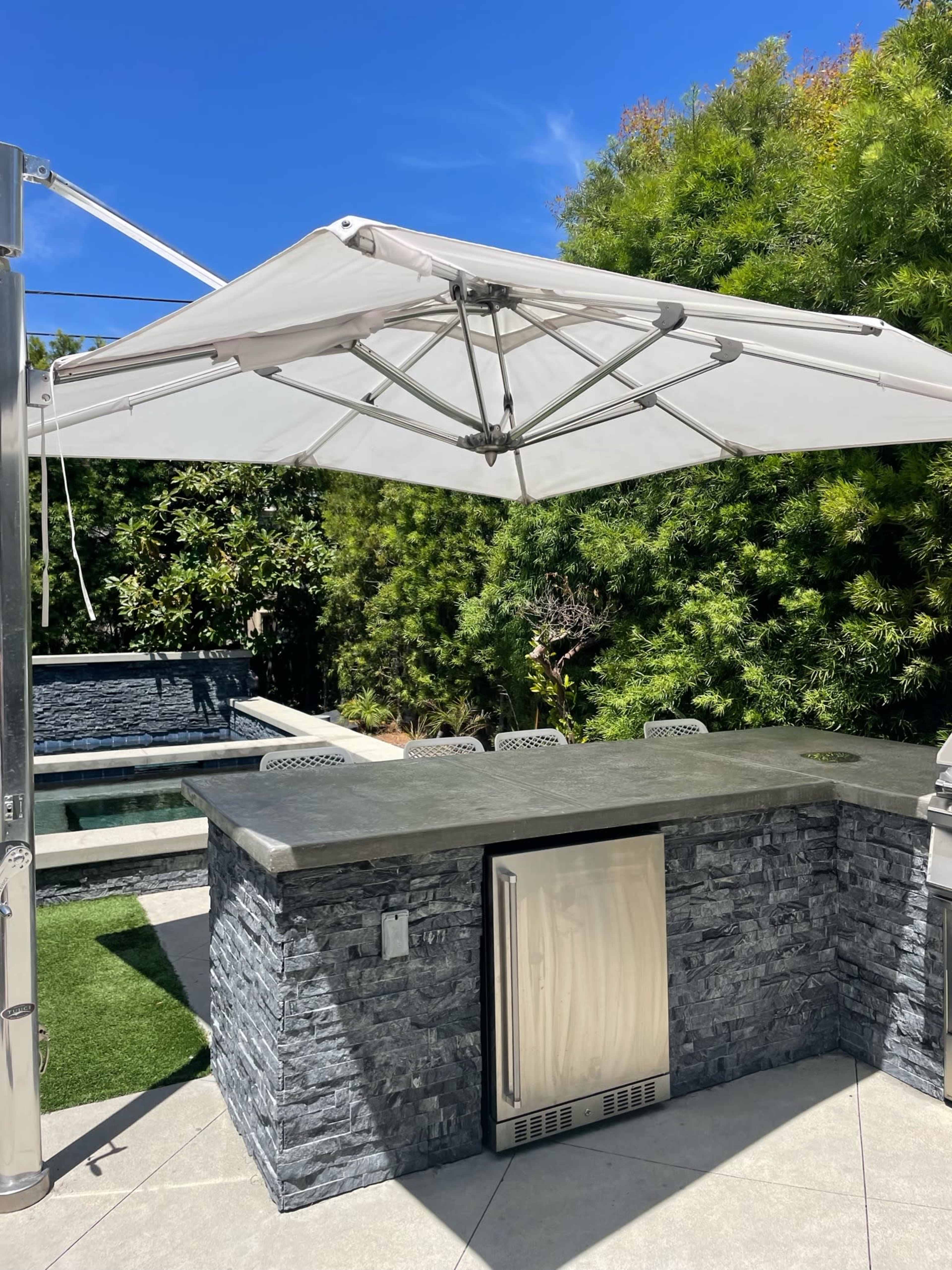 An outdoor kitchen area featuring a stone bar with a stainless steel refrigerator and a large white umbrella overhead, surrounded by greenery and a pool in the background.