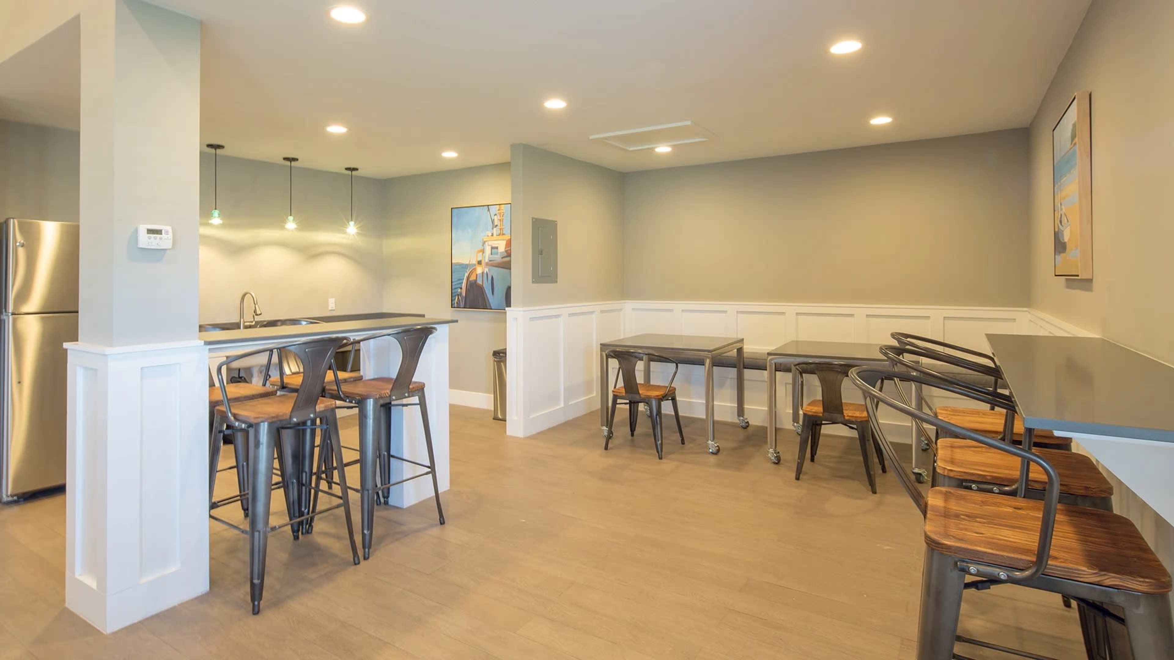 A modern kitchen and dining area with a bar counter, several tables, and chairs against a light-colored wall.