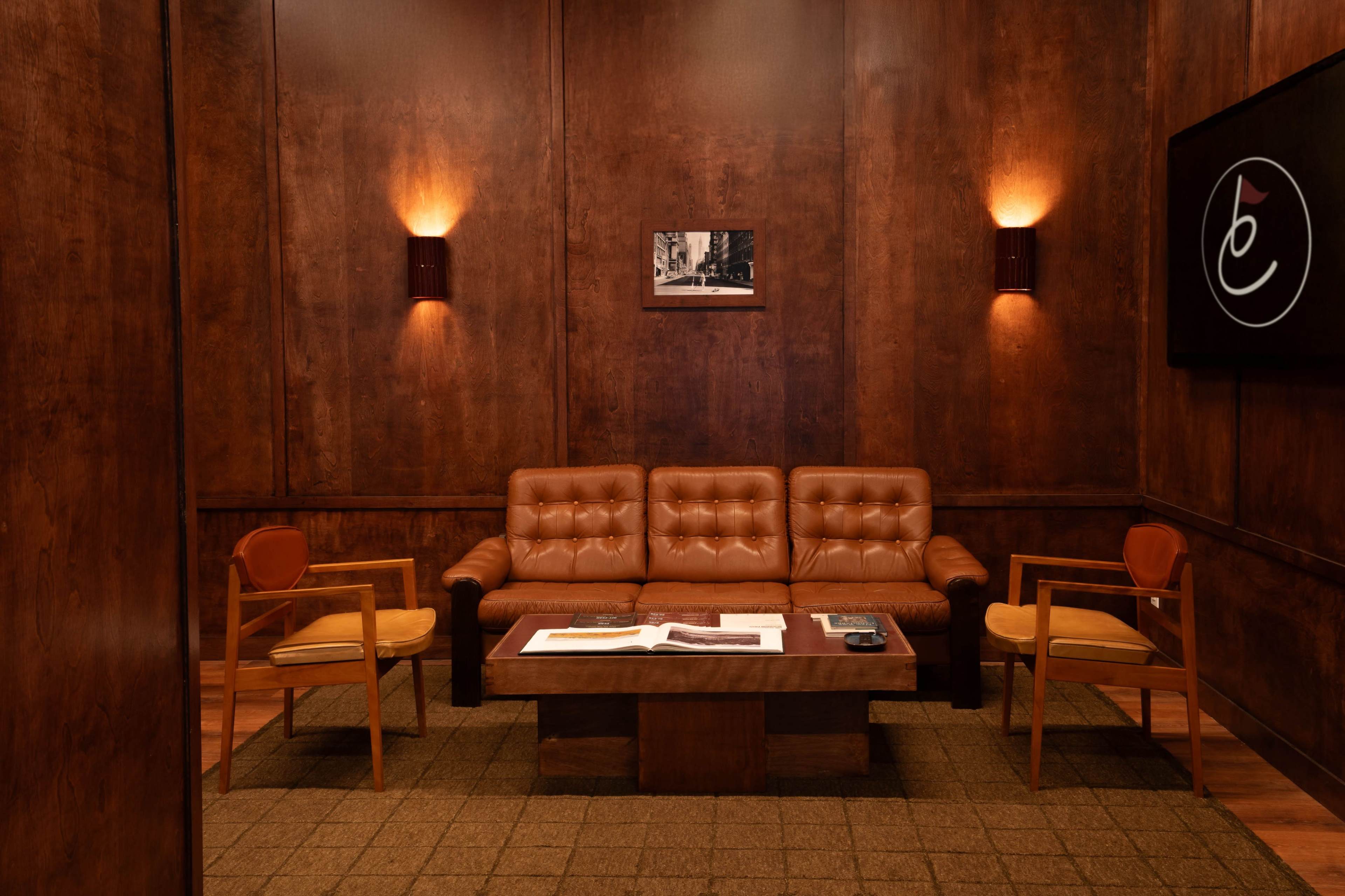 The image shows a wood-paneled room featuring a brown leather sofa, two wooden chairs, a low coffee table, and dim lighting.