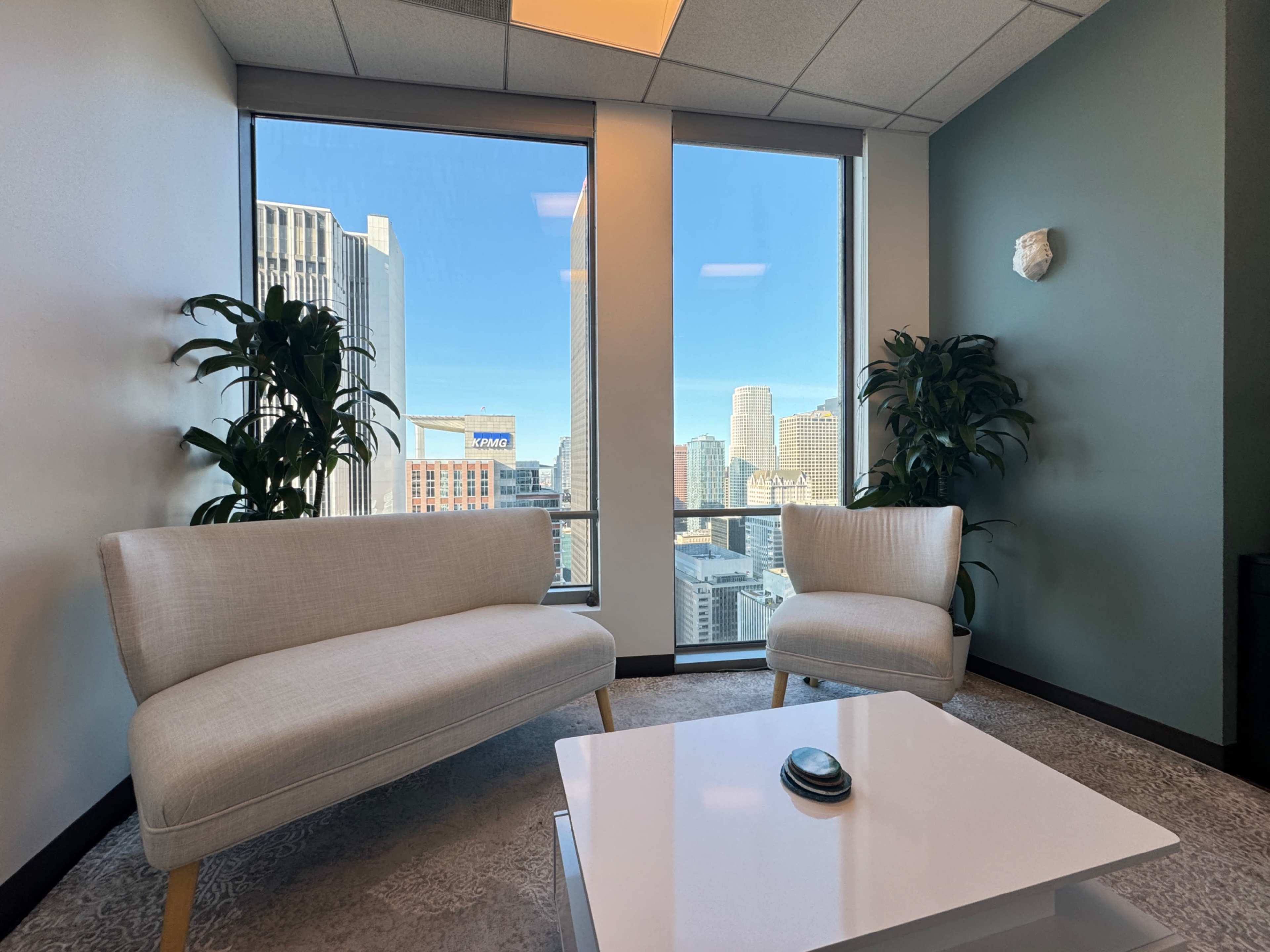A modern office lounge area features two light-colored sofas and a coffee table, with large windows showcasing a city skyline.