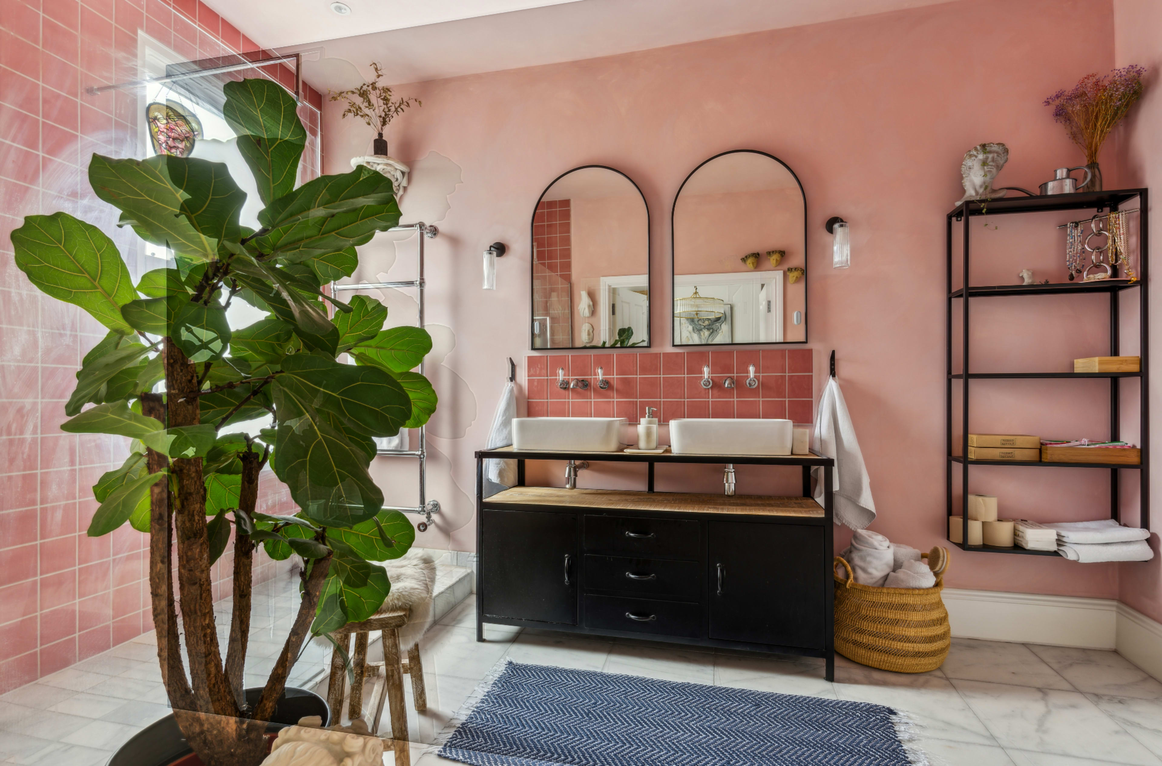 A modern bathroom features pink tiled walls, a black double sink vanity, two round mirrors, a large indoor plant, and stylish shelving.