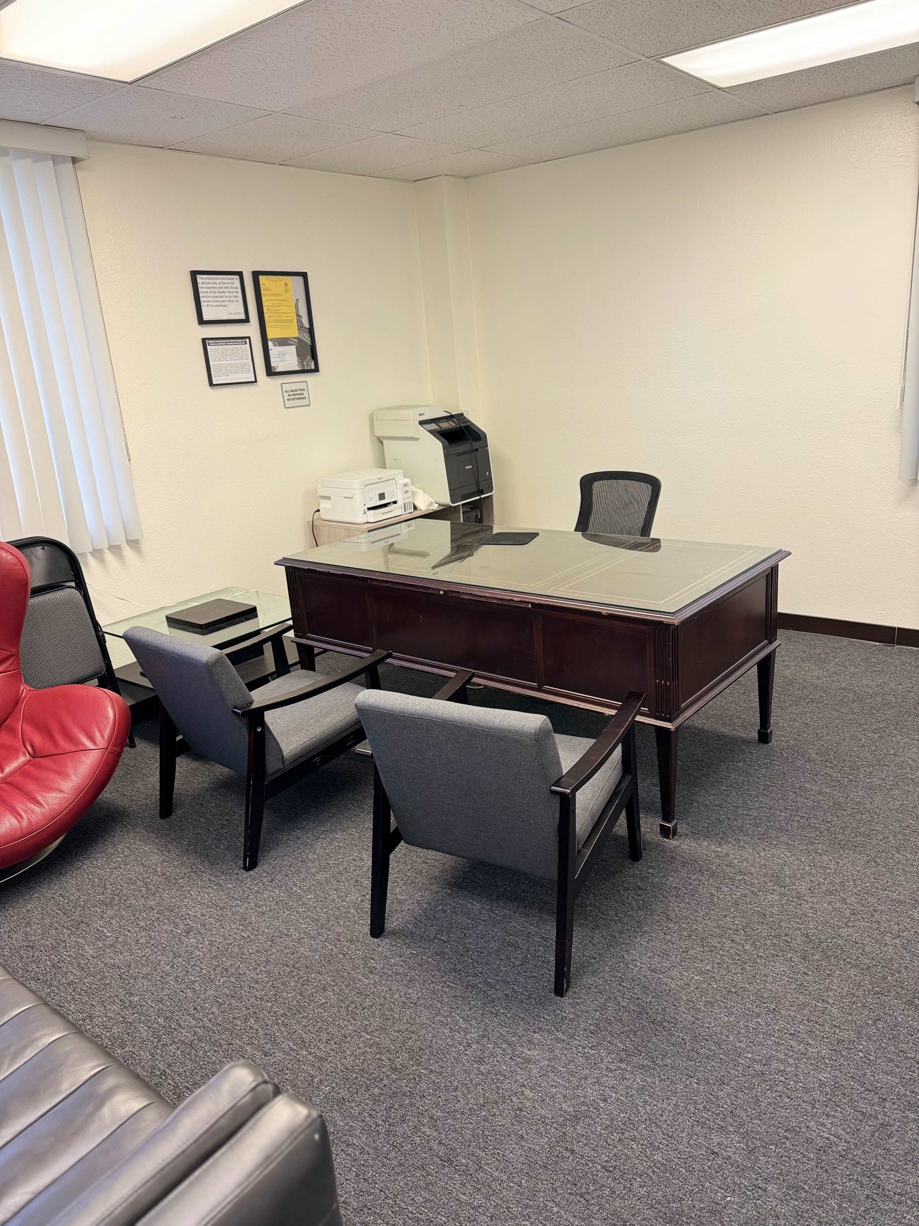 A simple office space with a wooden desk, two gray chairs, a red lounge chair, and a printer in the corner.