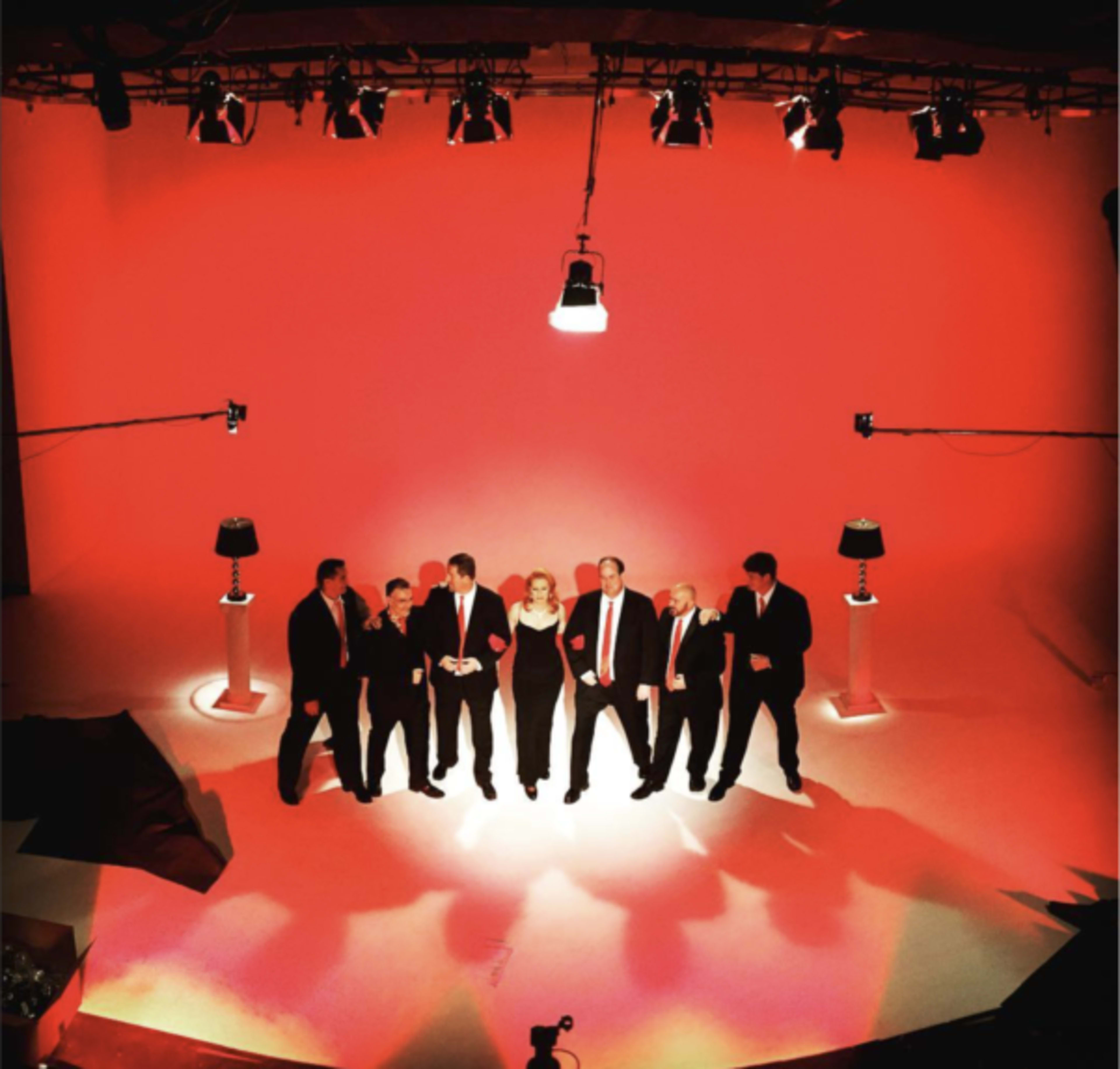 A group of eight individuals in formal attire stands together on a brightly lit stage with red walls and two lamps.