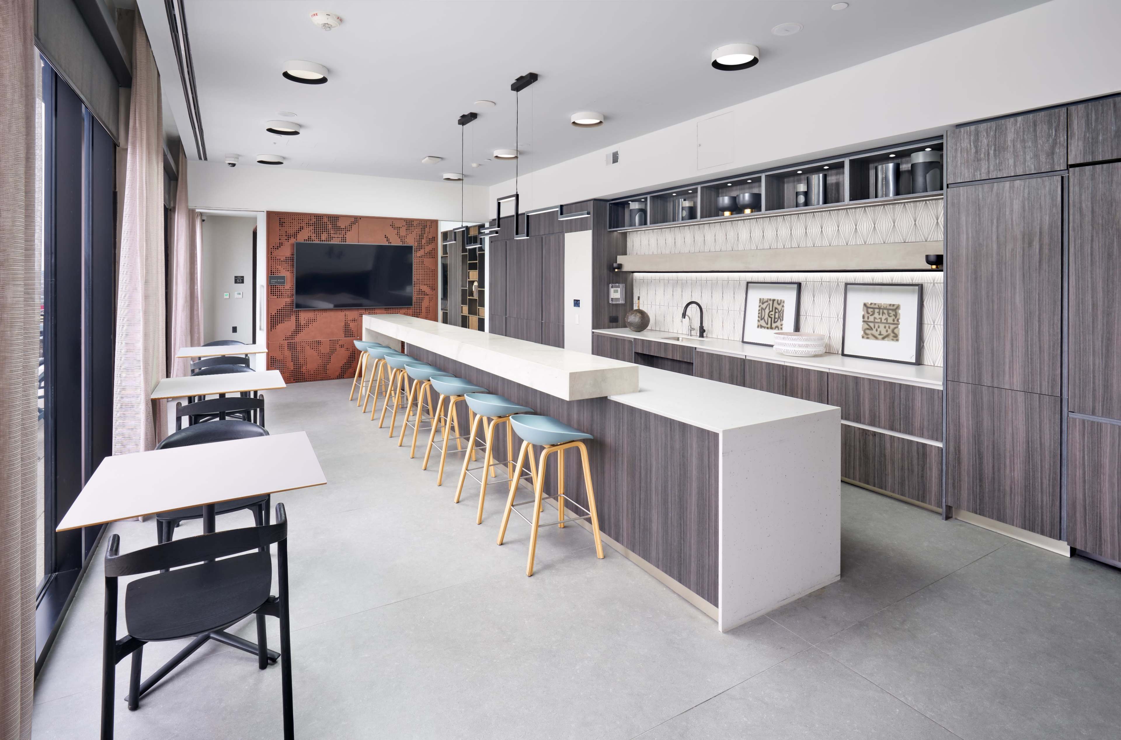 The image shows a modern kitchen area featuring a long, light-colored countertop with bar seating and wooden cabinets, alongside a wall-mounted television and a large window.
