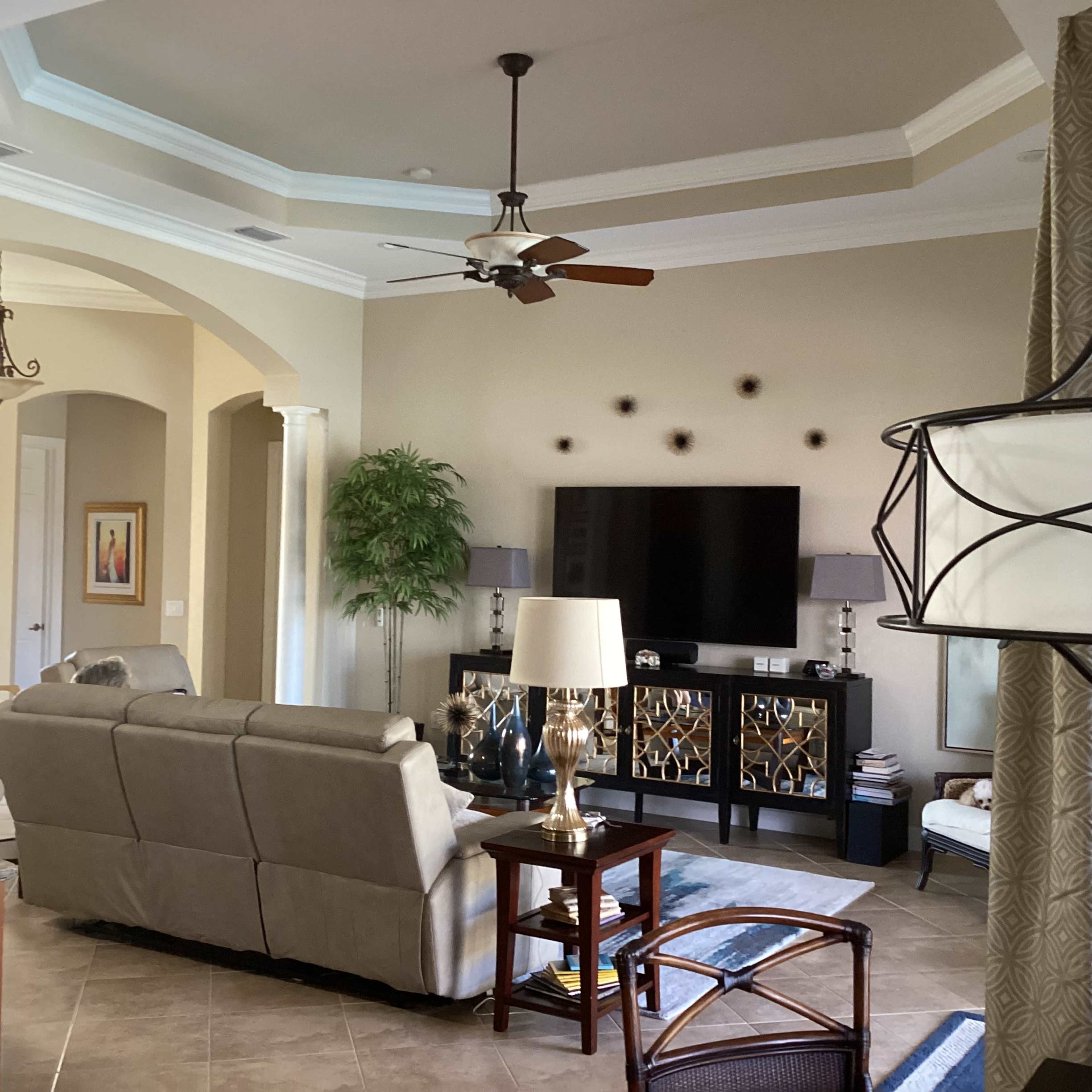 A modern living room featuring a beige sectional sofa, a coffee table, and a TV cabinet against a light-colored wall, with a ceiling fan and potted plant in the background.