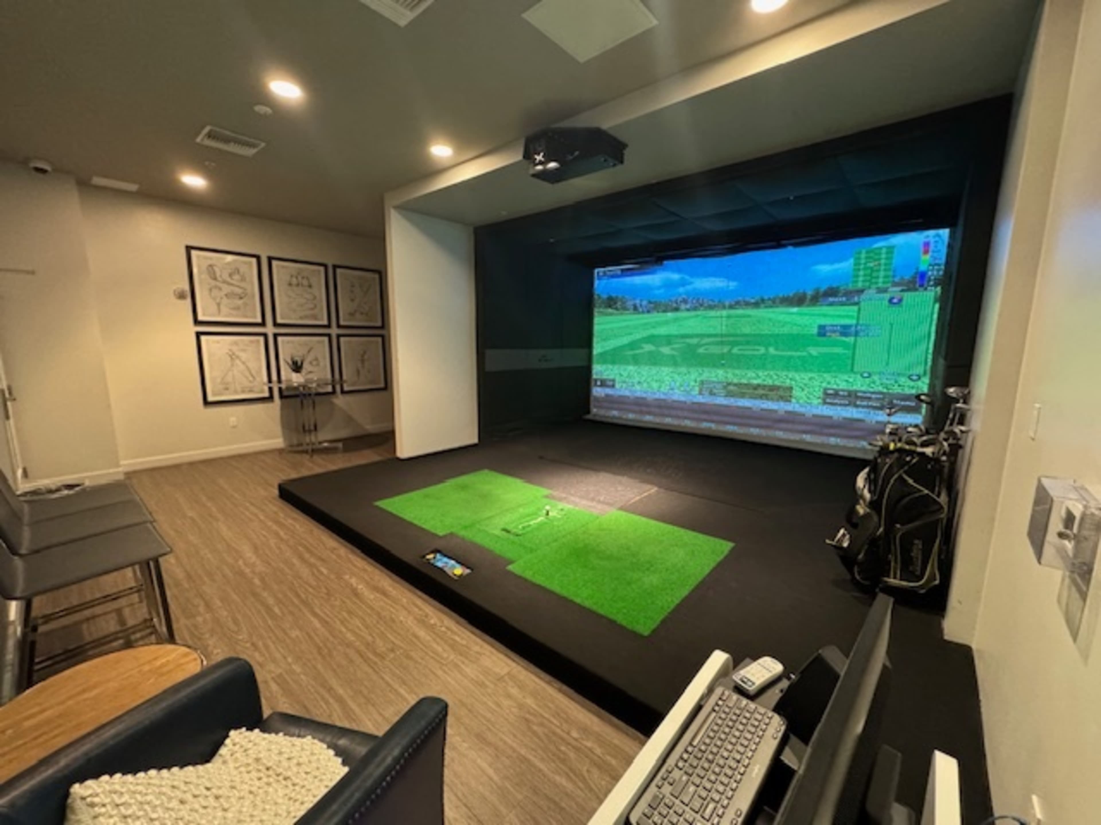 The image shows an indoor golf simulation room featuring a large screen displaying a golf course, synthetic grass areas for putting, and golf clubs nearby.