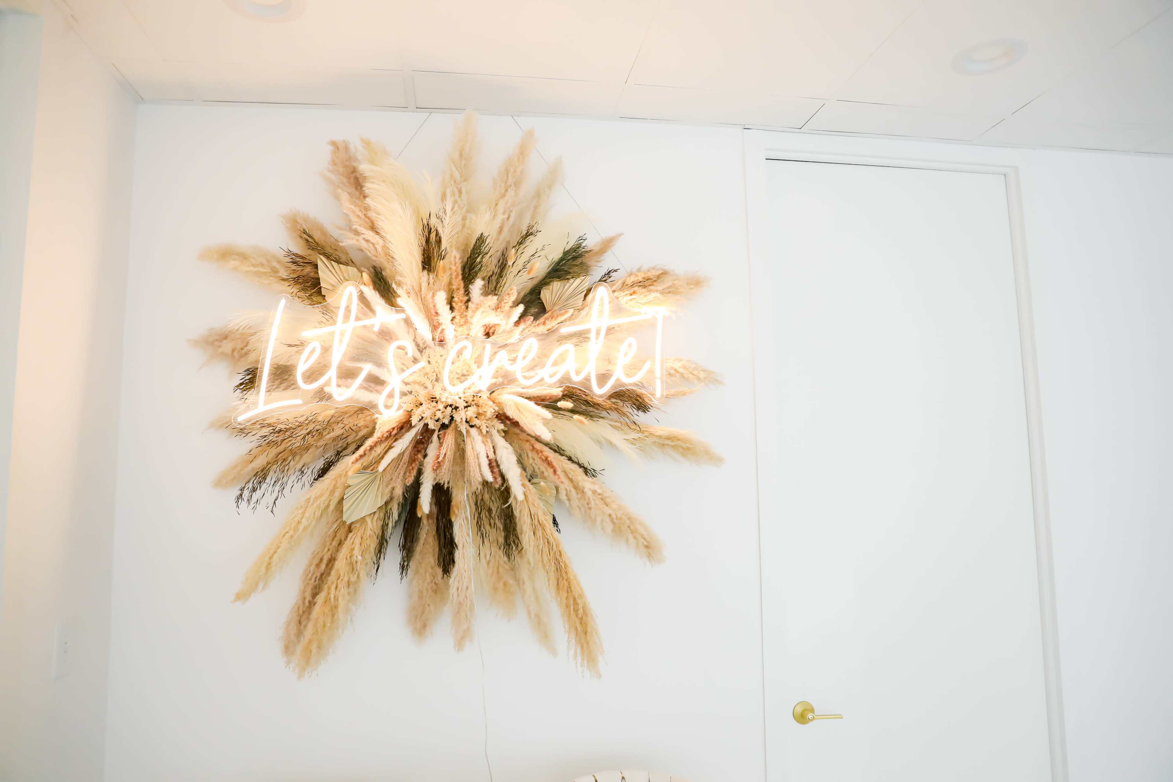 A decorative wall piece featuring dried plants and a neon sign that reads "Let's create" is displayed against a white wall.
