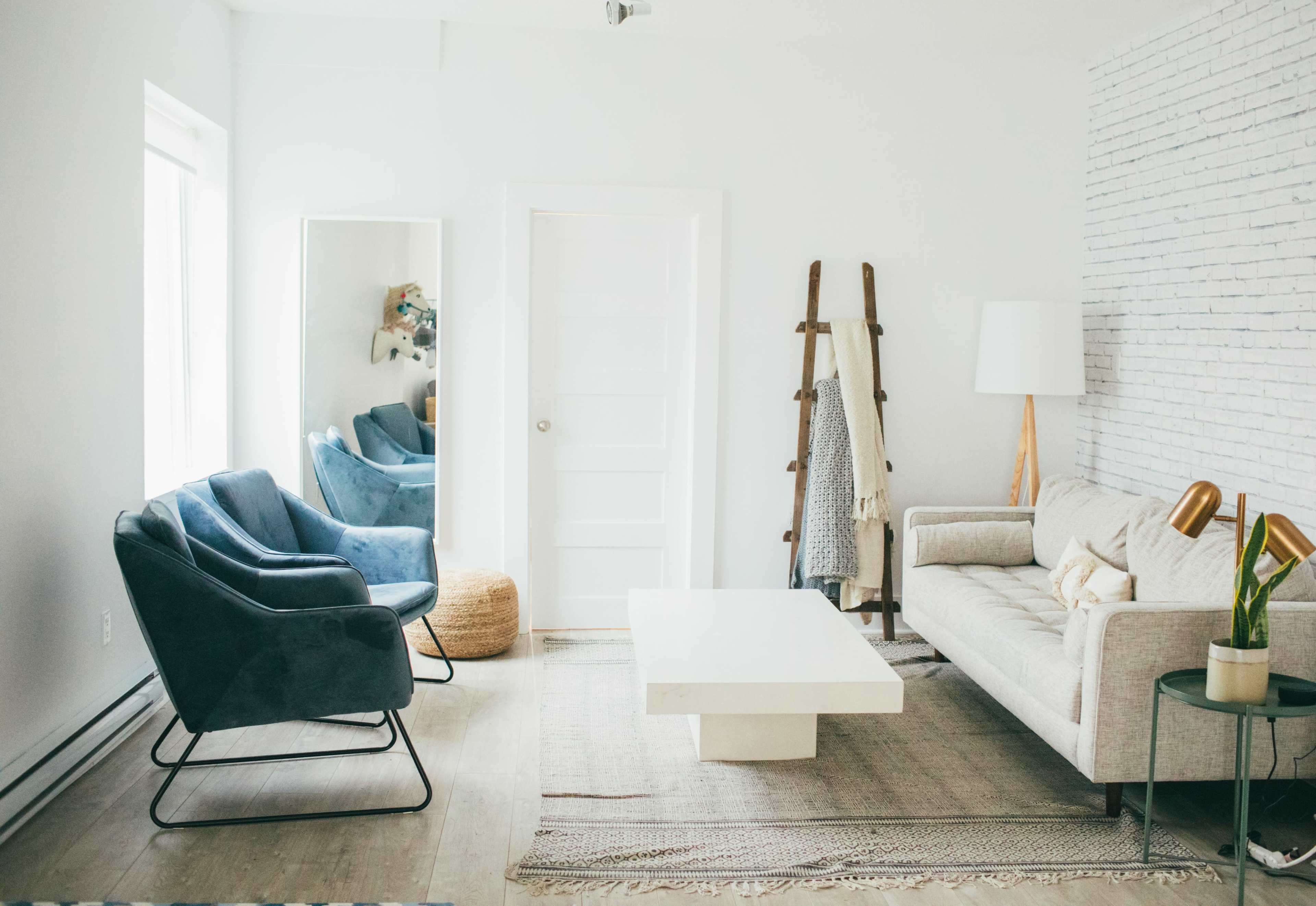 A bright living room features a gray sofa, two blue armchairs, a large white coffee table, and a floor lamp, complemented by a mirror and a decorative ladder against the wall.