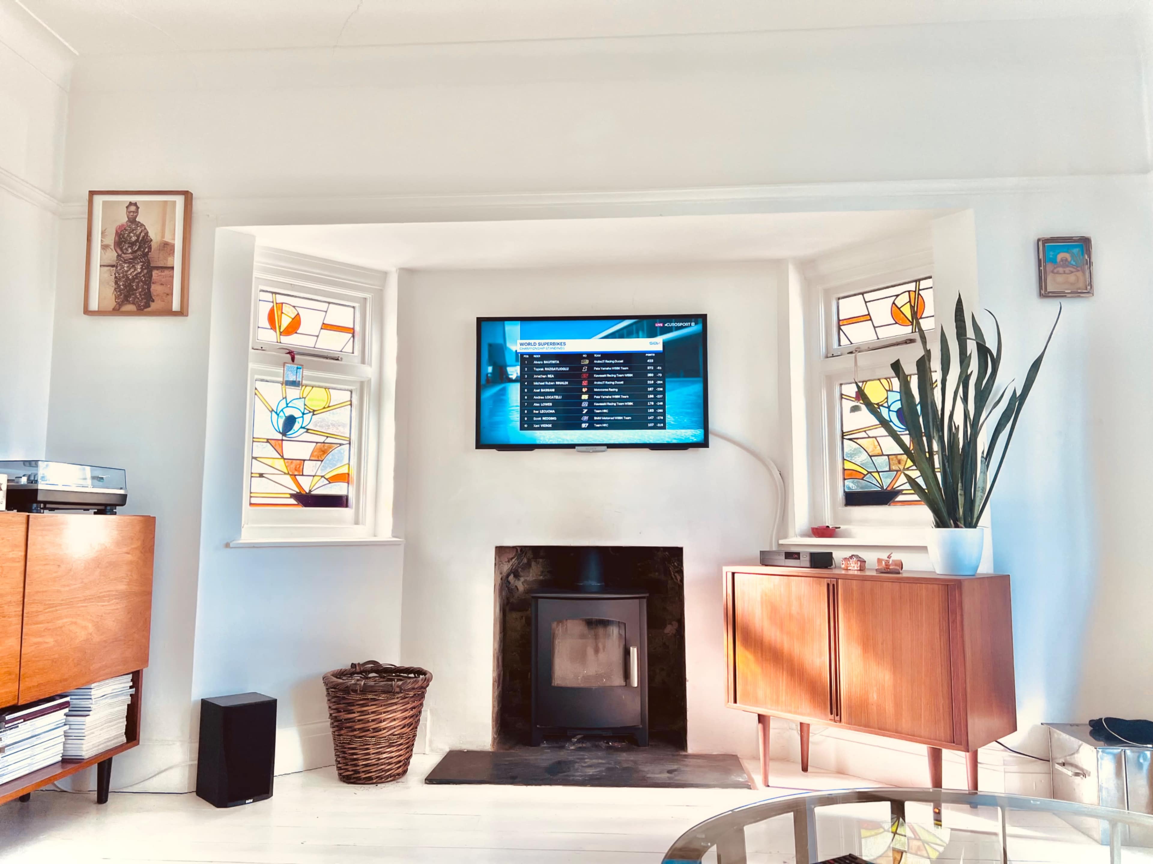 A living room with a TV mounted on the wall above a modern fireplace, flanked by stained glass windows, a wooden cabinet, and a decorative plant.
