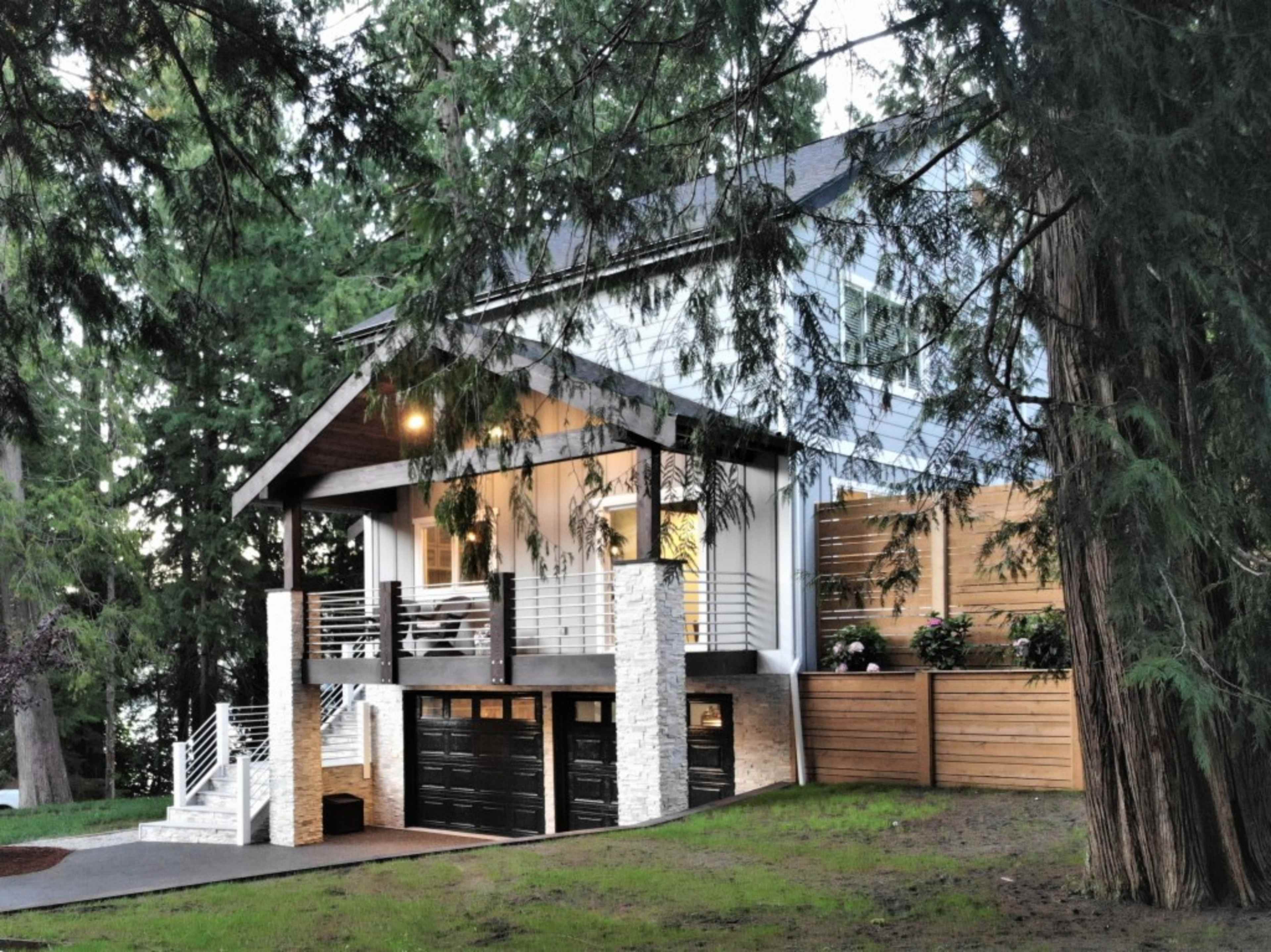 A modern house with a covered porch and two garage doors is surrounded by tall trees.