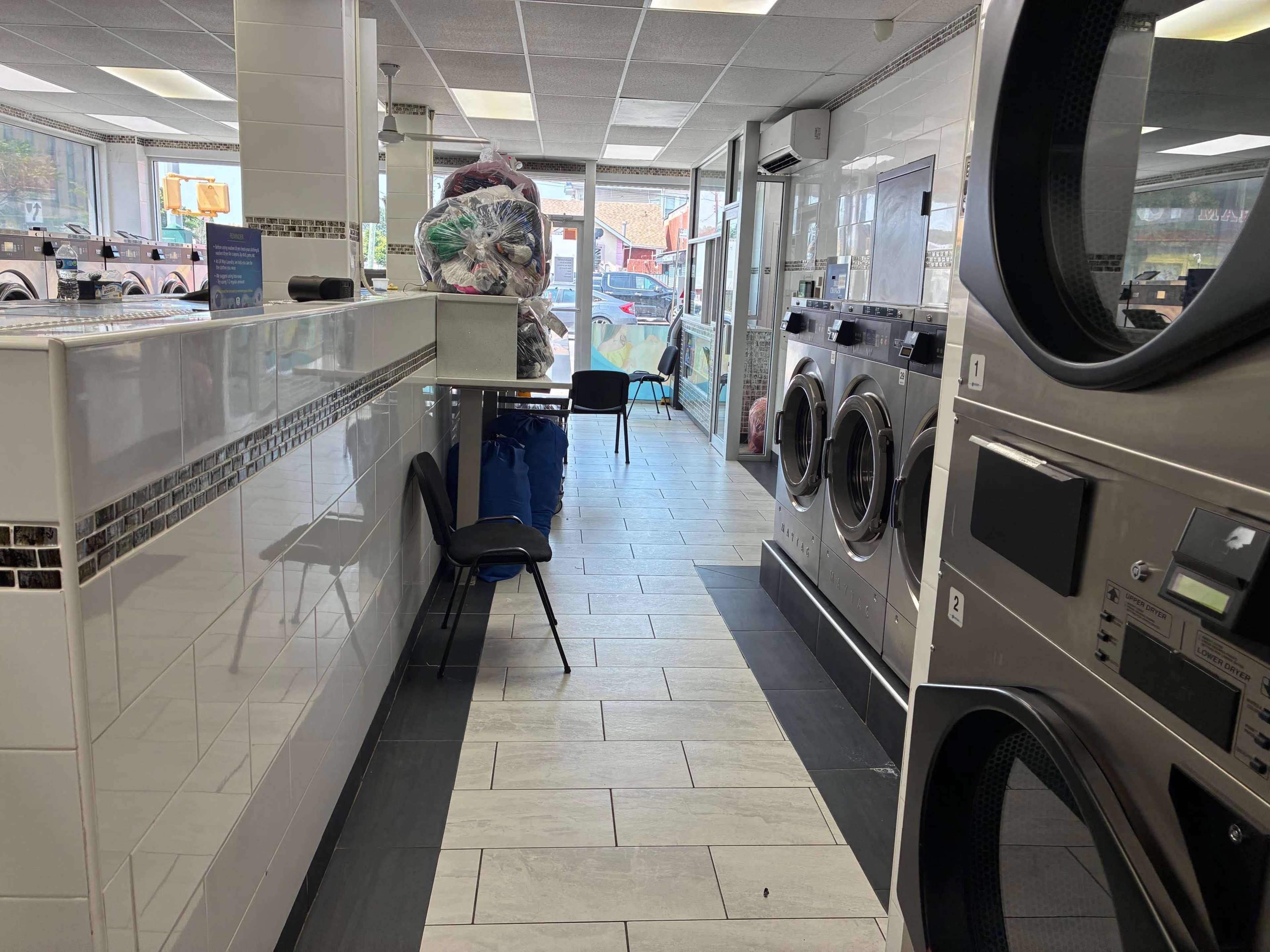 The image shows the interior of a laundromat with rows of washing machines, a seating area, and a view of the outdoors through large windows.