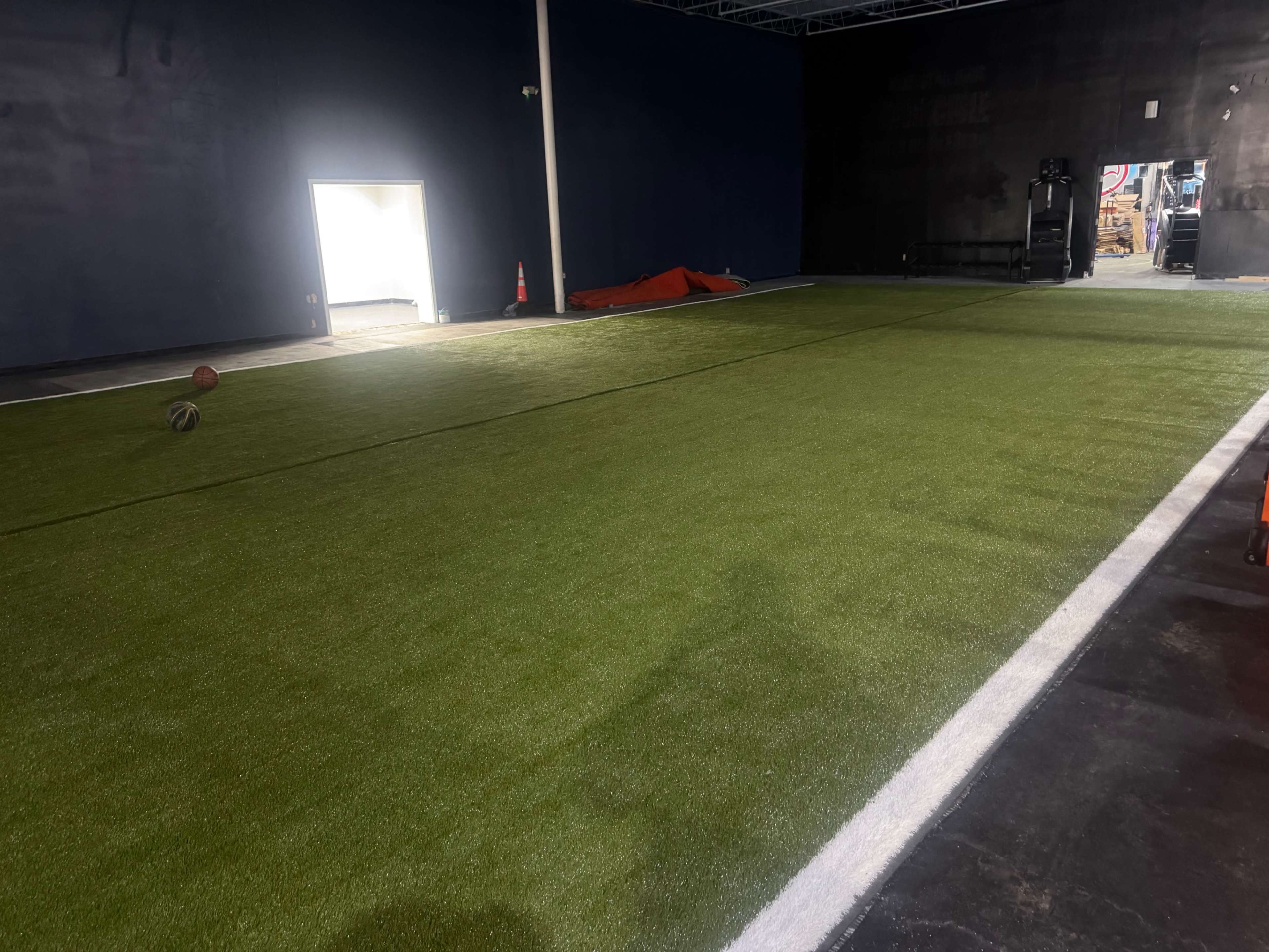 The image shows an indoor space with a green artificial turf surface and a single open door letting in light, while a basketball and some equipment are visible on one side.