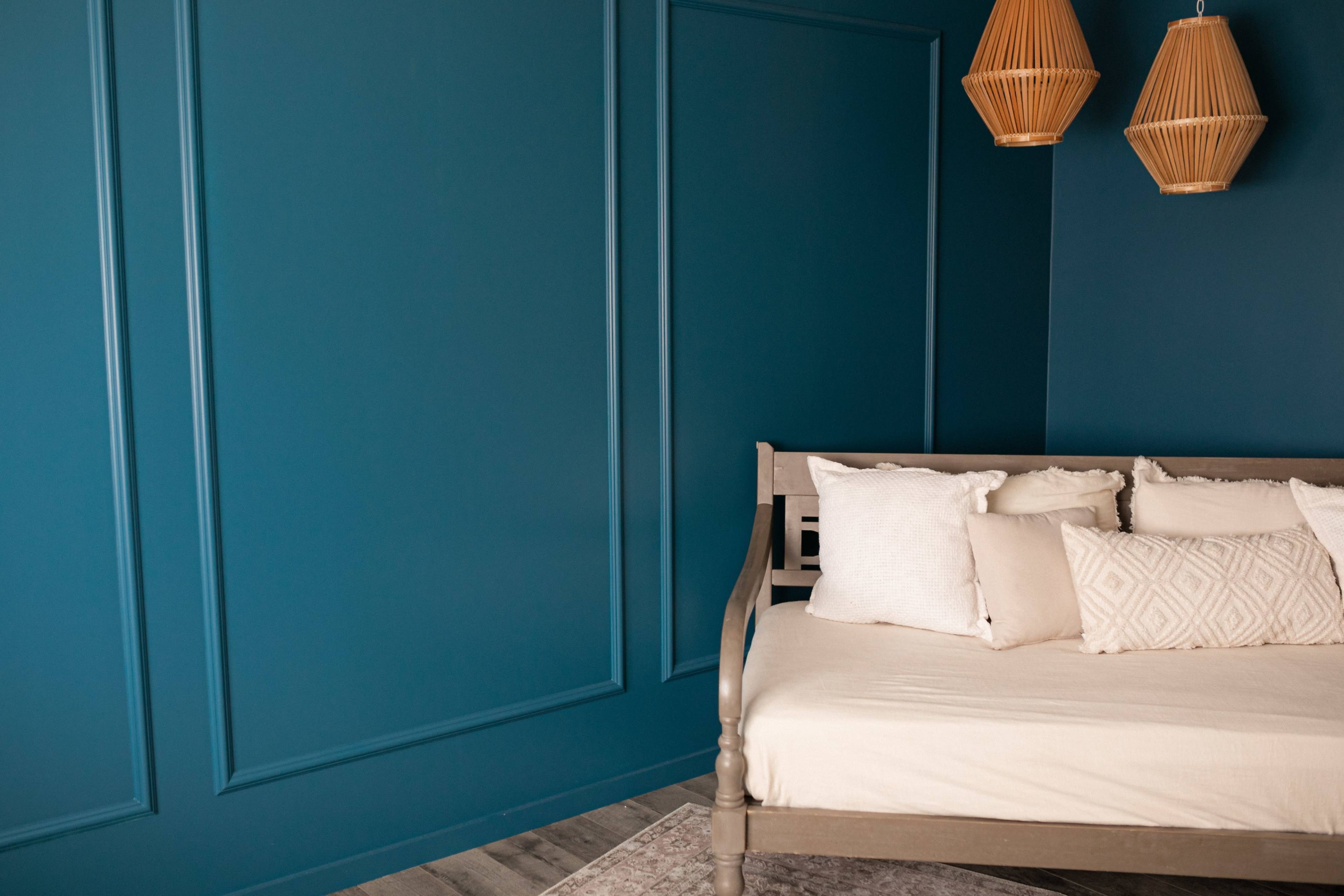 A corner of a room featuring a daybed with cream-colored pillows against a dark blue paneled wall and two woven pendant lights hanging above.