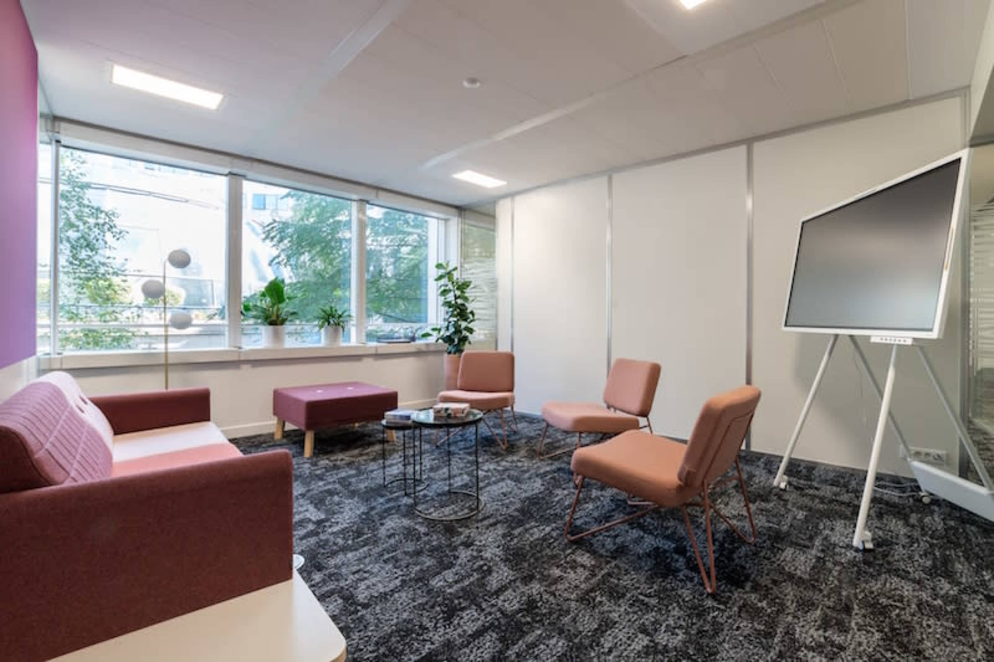 A modern office lounge area featuring a pink and white seating arrangement, a coffee table, and a large screen on a stand, surrounded by windows and plants.