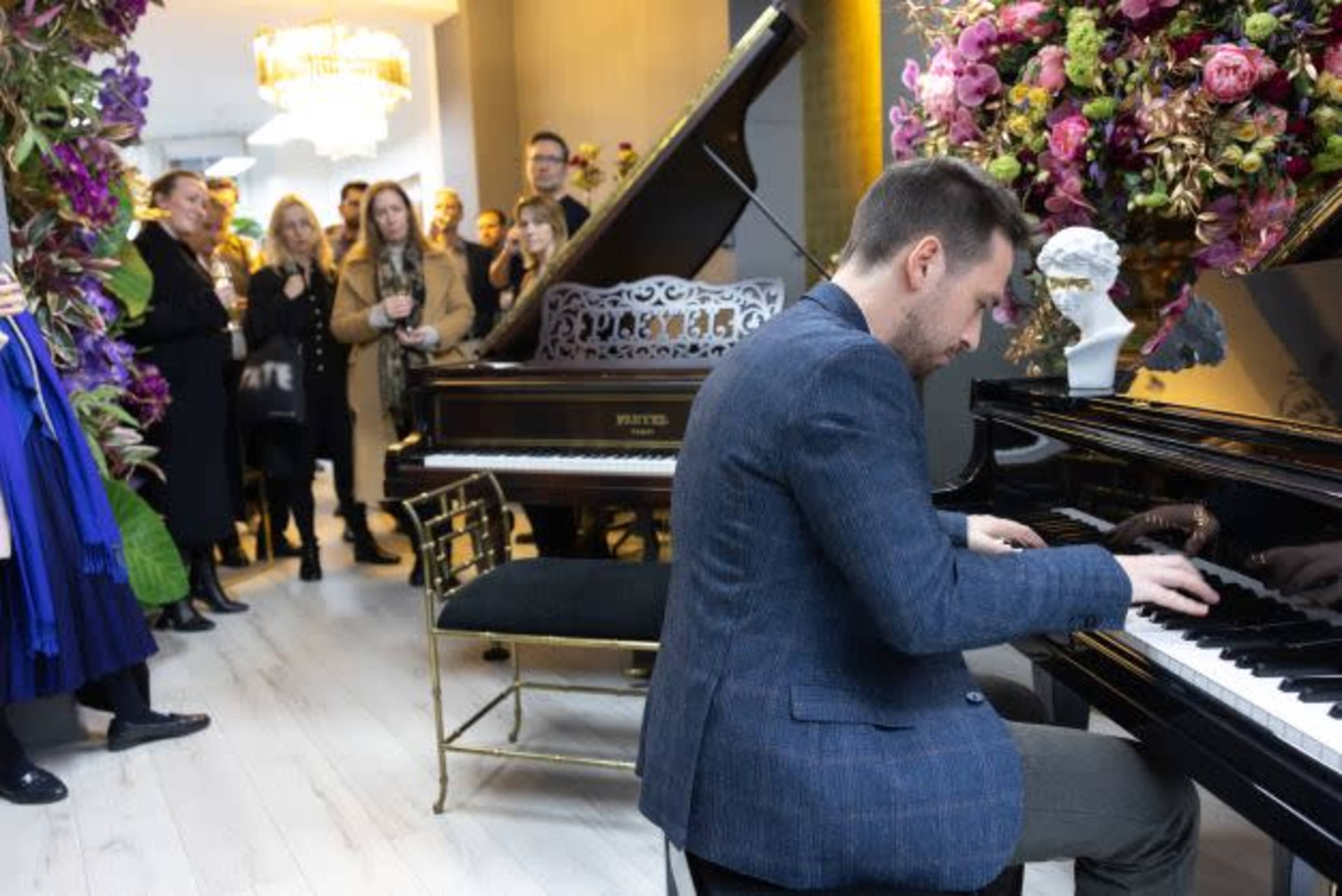 A man plays a grand piano in front of an audience surrounded by flower arrangements and elegant decor.