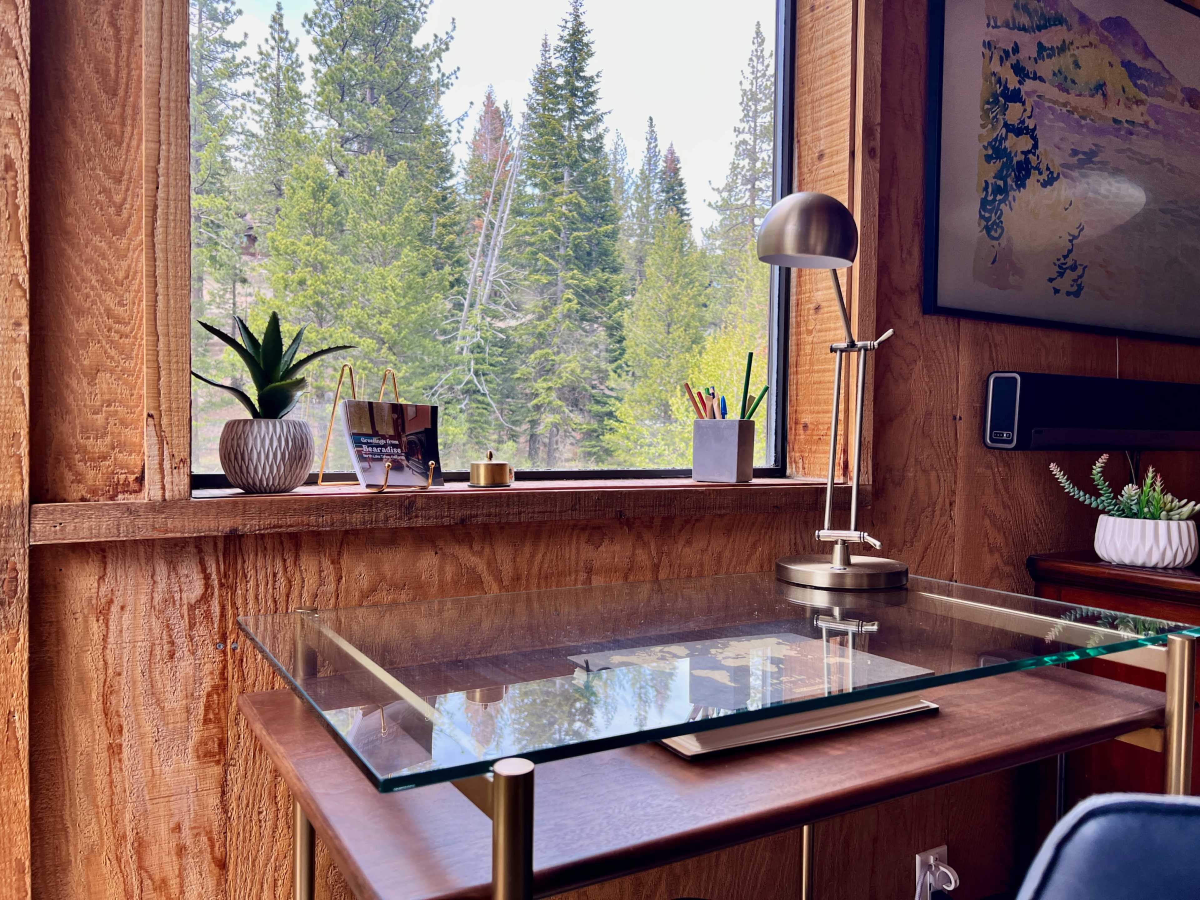 A modern desk with a glass top is positioned near a window overlooking a forested landscape, featuring plants and stationery on the surface.