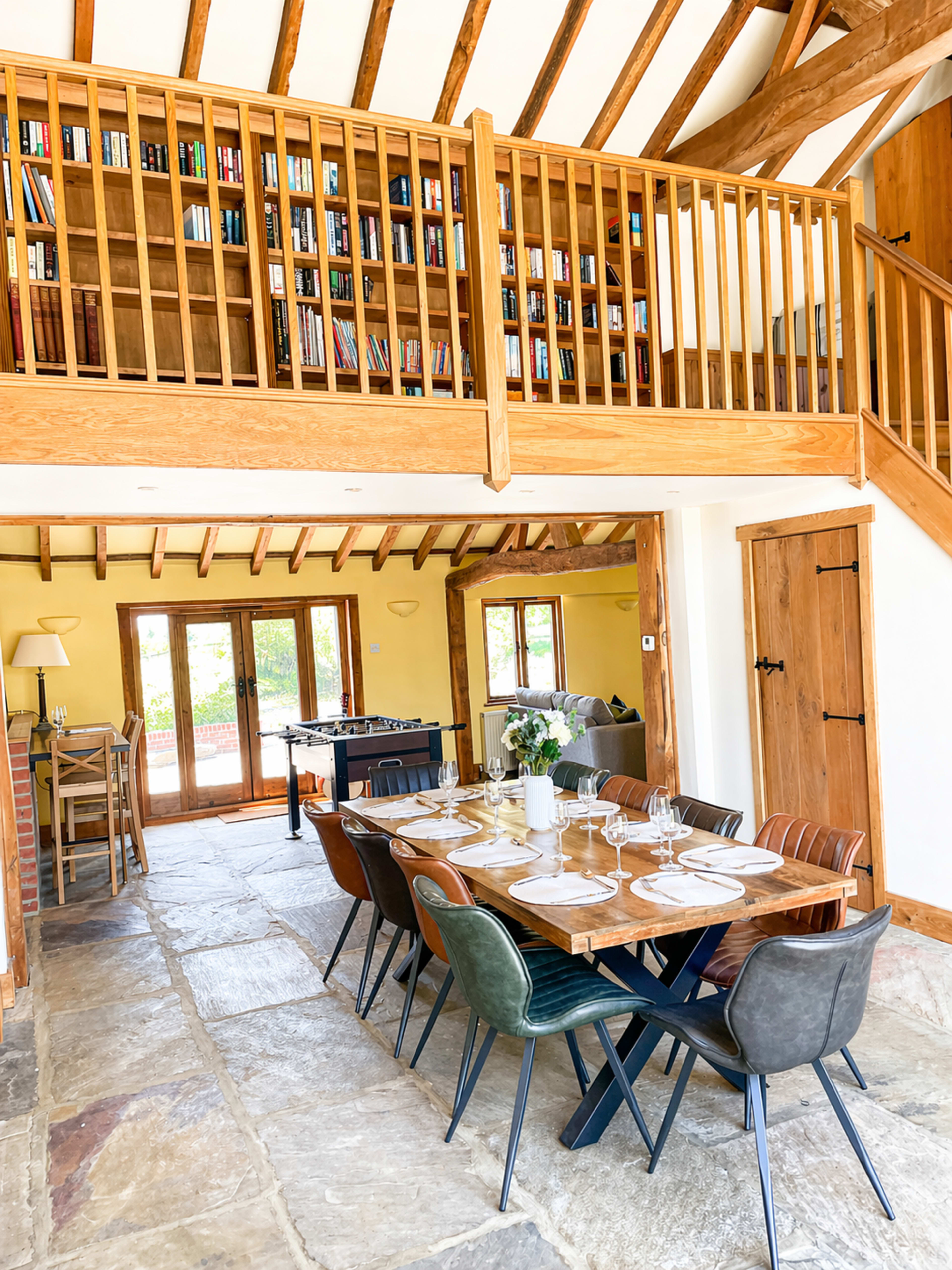 The image shows a spacious dining area with a long table set for a meal, featuring a wooden staircase and a loft with bookshelves in the background.