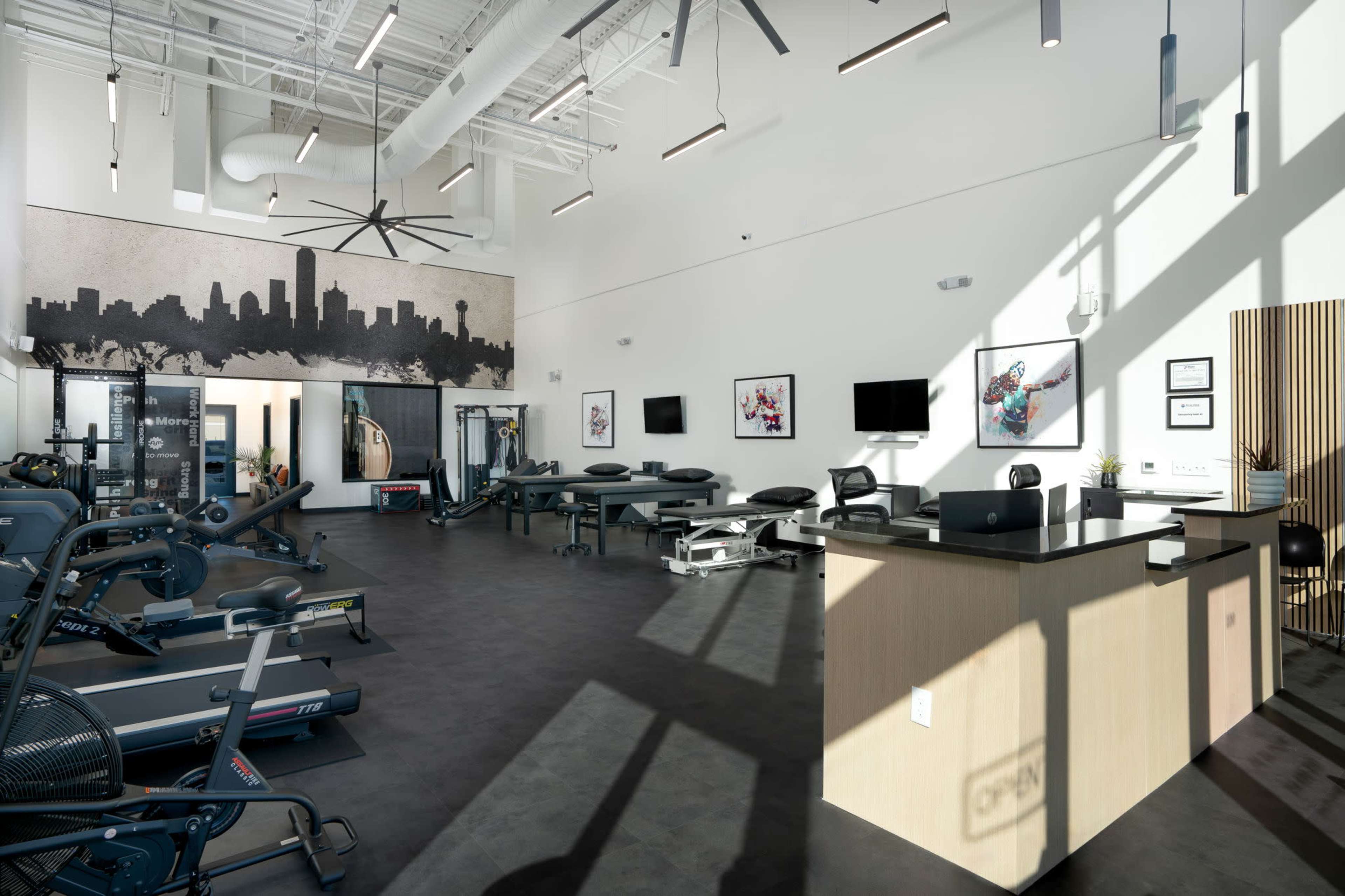 The image shows a modern fitness facility featuring exercise equipment, a reception area with a counter, and large windows allowing natural light to enter.