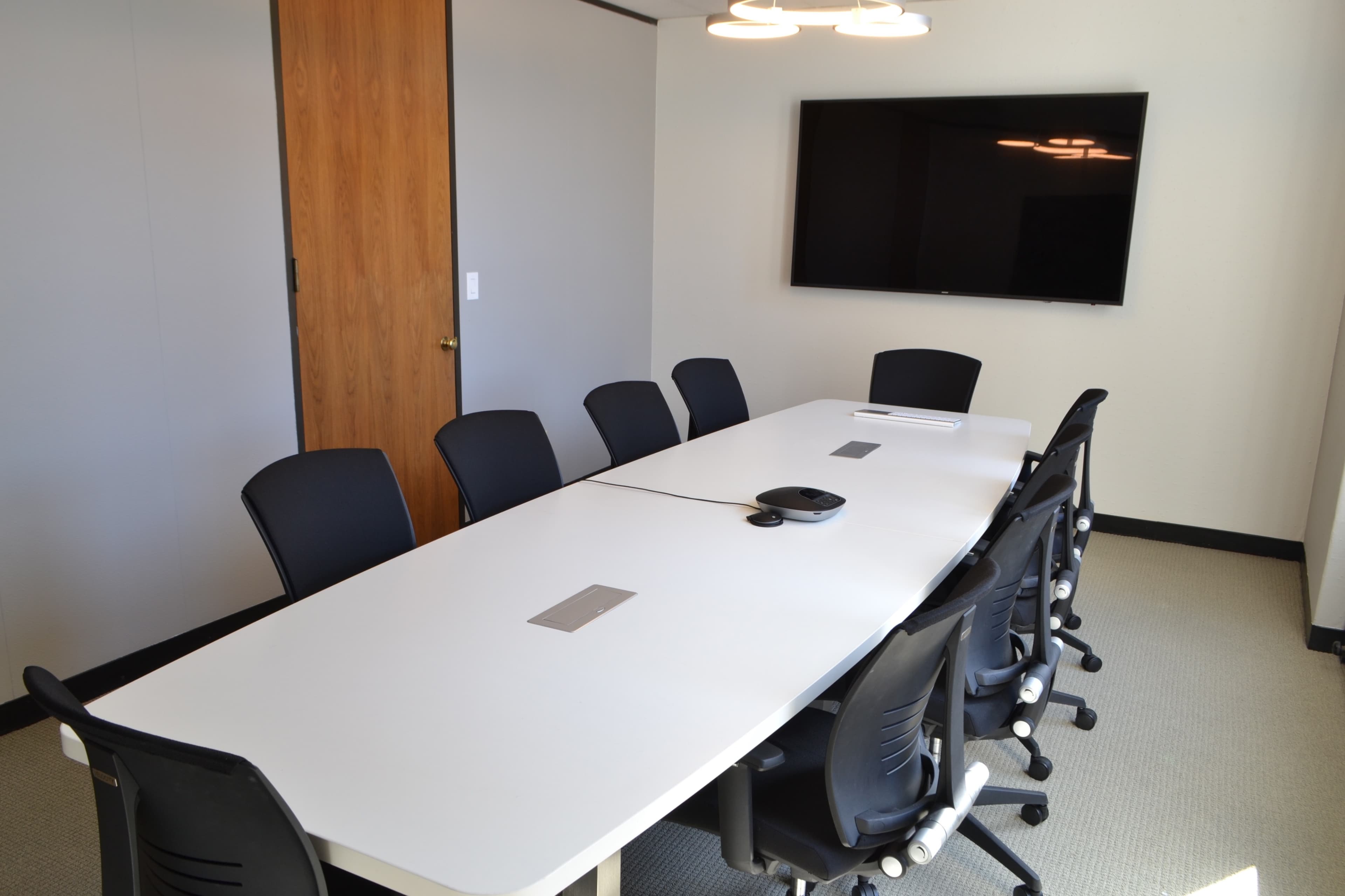 A modern conference room features a long white table surrounded by black chairs and a wall-mounted television.