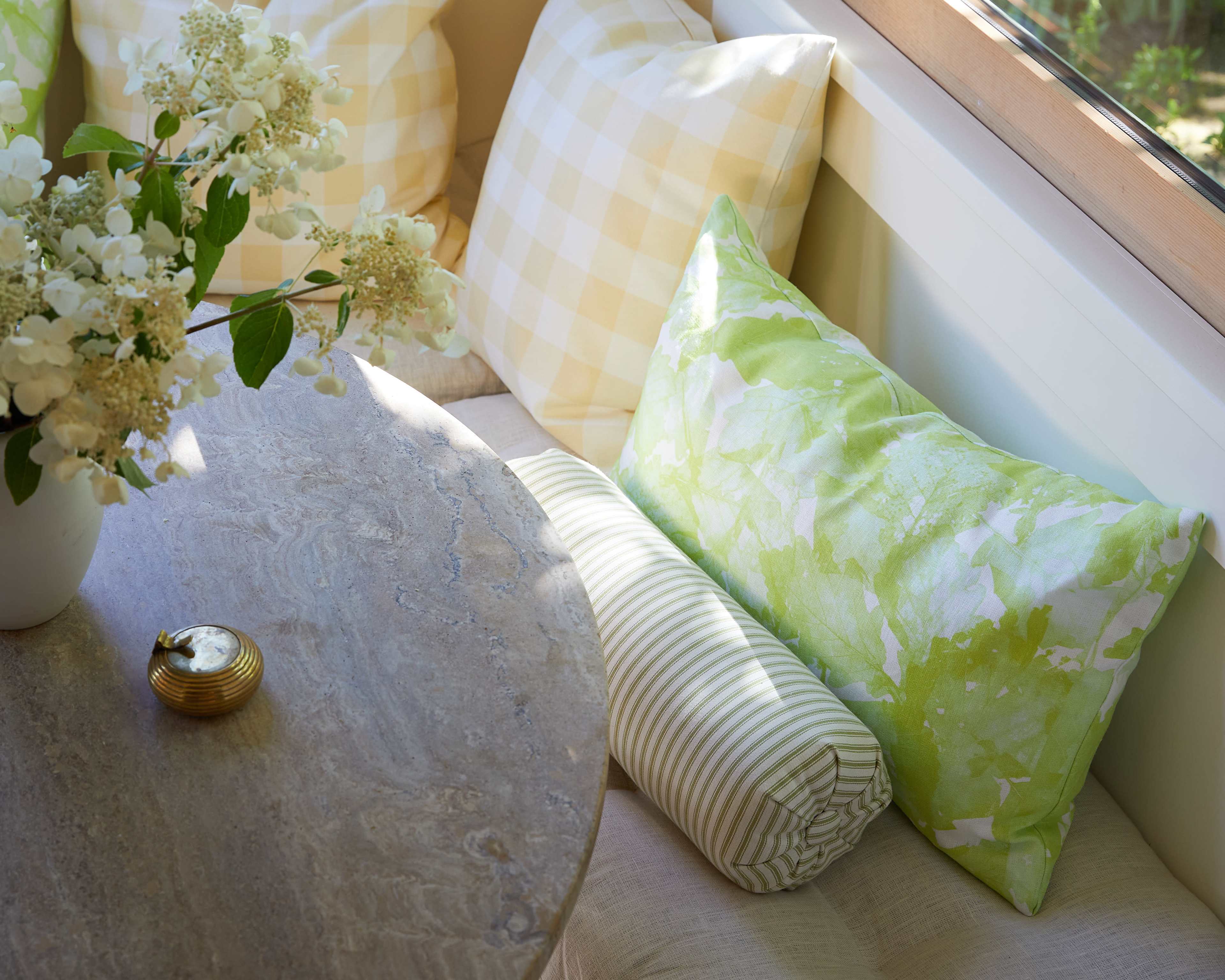 A cozy seating area features a round stone table with decorative pillows in various patterns and colors, accompanied by a vase of flowers.