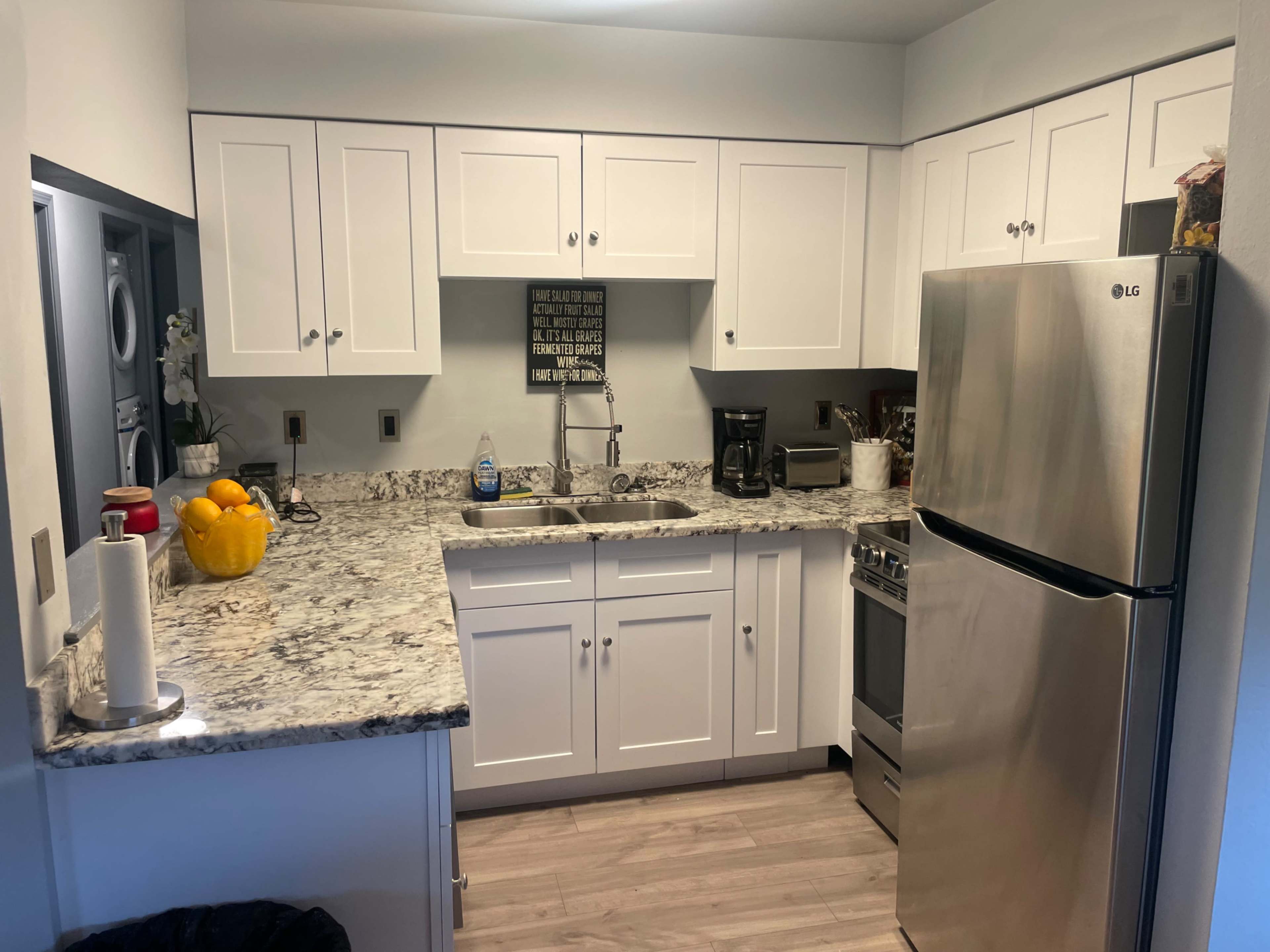 A modern kitchen features white cabinets, a granite countertop, stainless steel appliances, and a bowl of oranges on the counter.