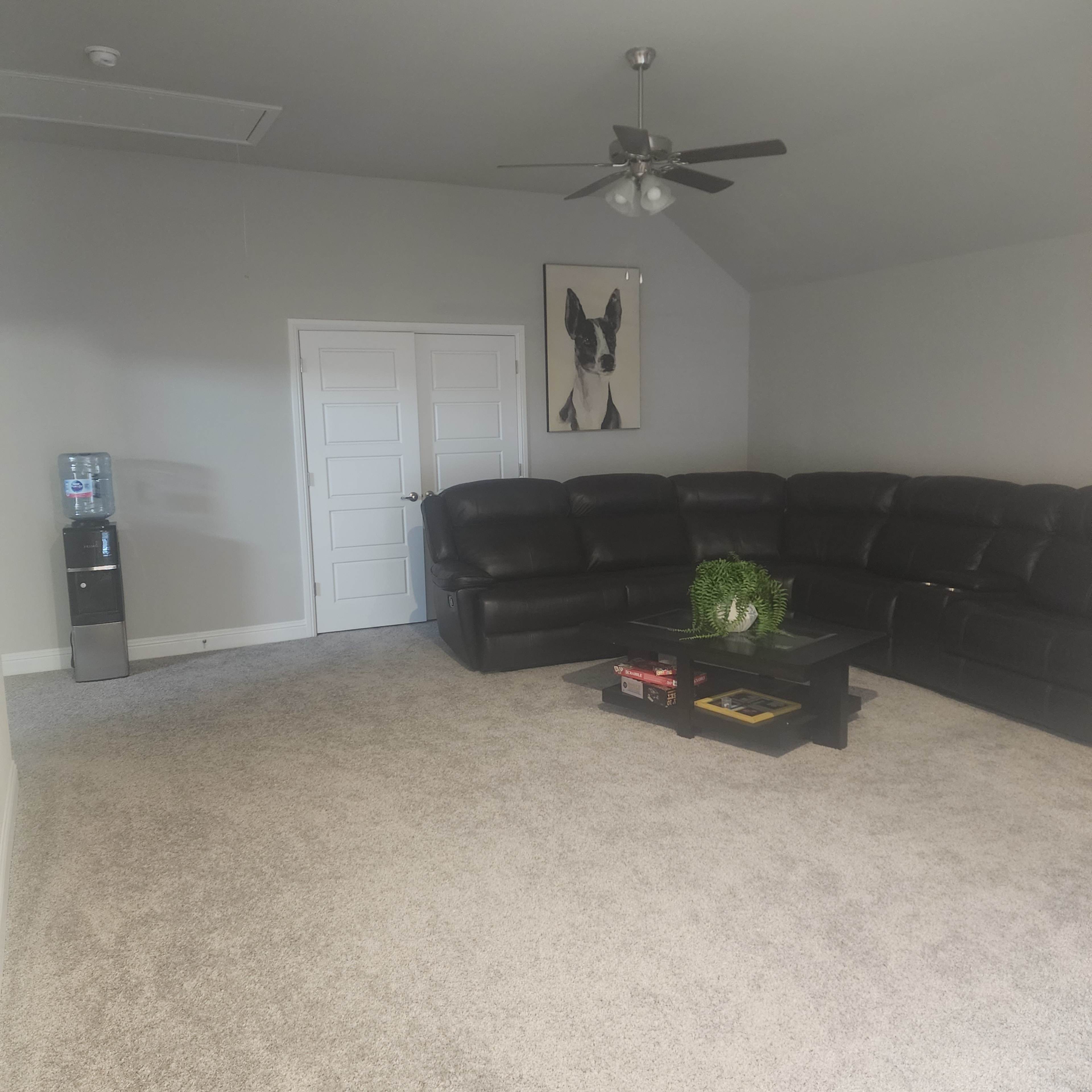 A spacious room features a black leather sectional sofa, a coffee table with a plant, a water cooler, and a framed picture on the wall.