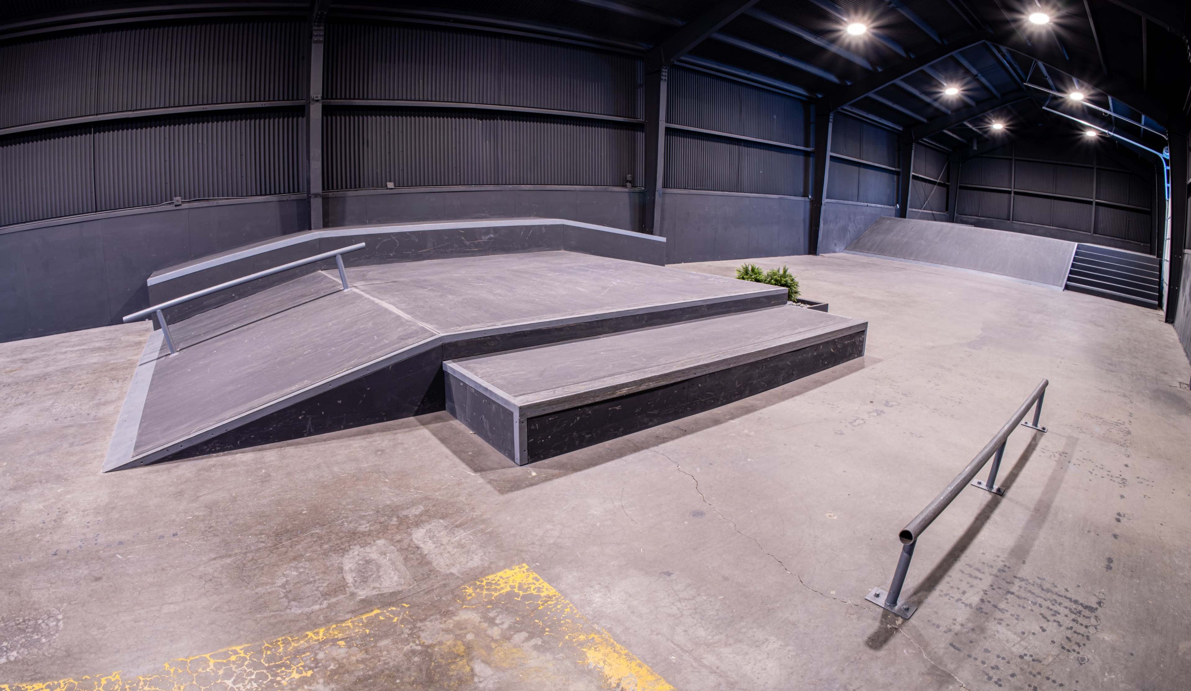 The image shows an indoor skateboard park featuring various ramps and a rail in a spacious warehouse setting.