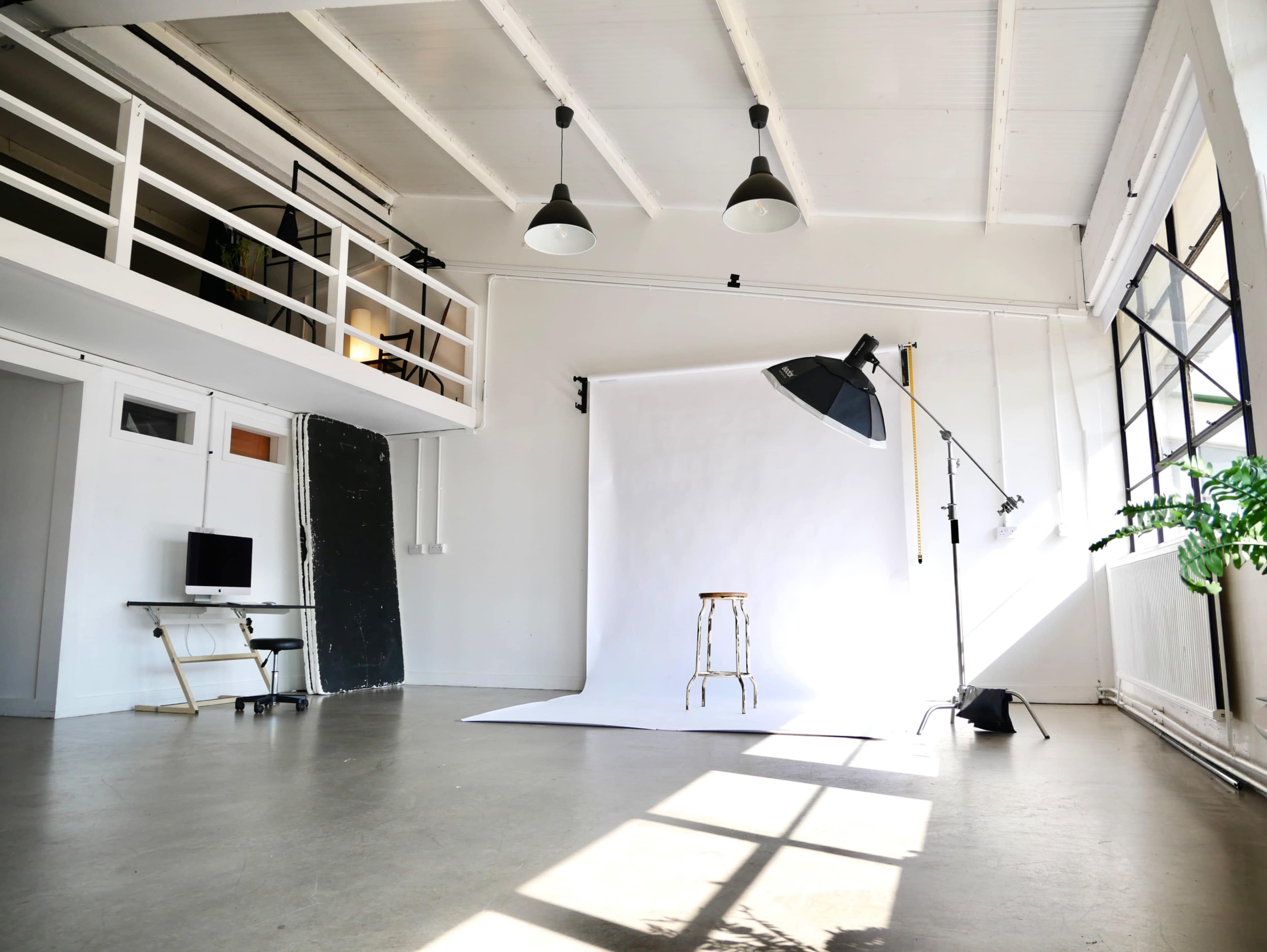 The image shows a spacious, well-lit photography studio with a white background, a stool, and studio lighting equipment.