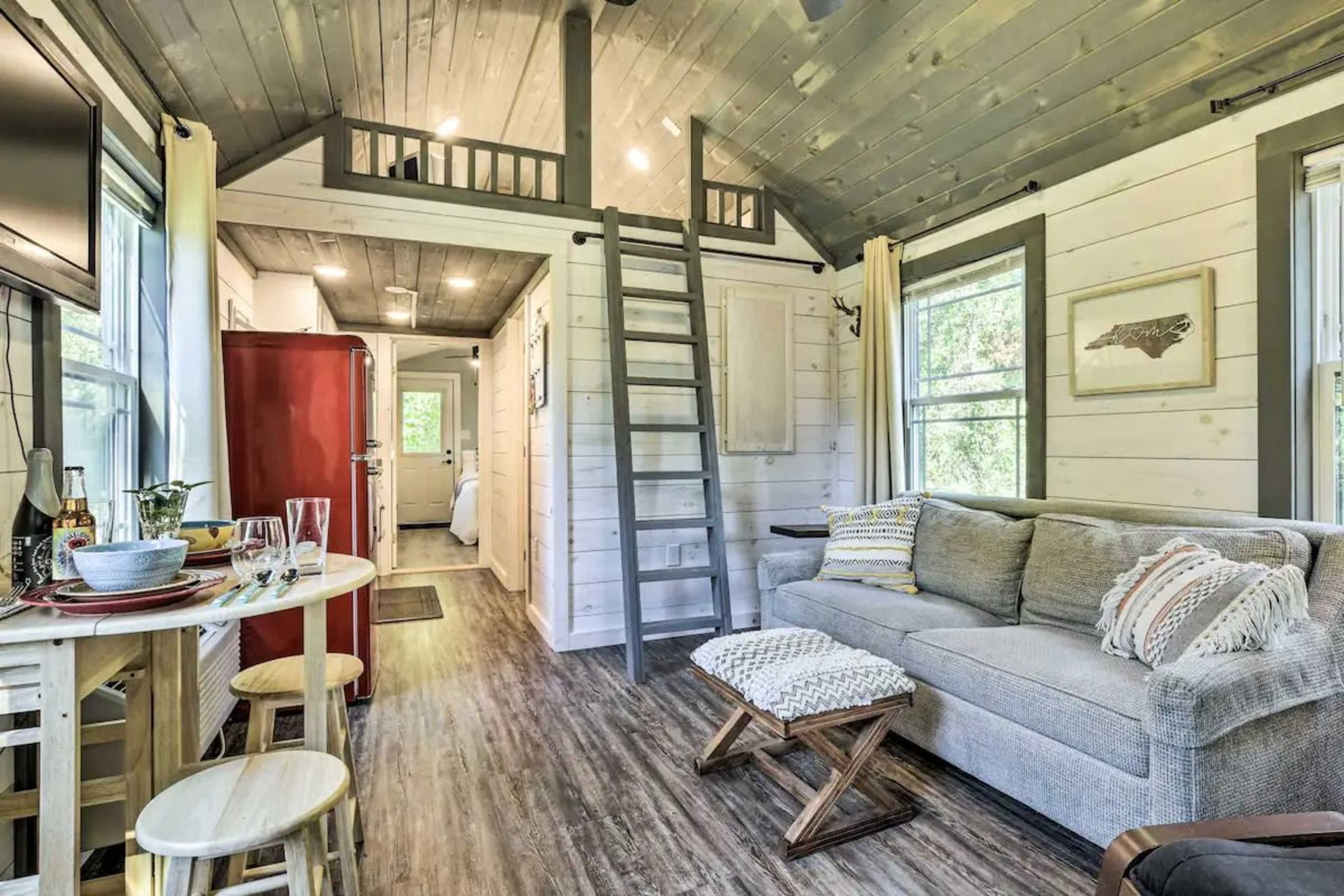 A cozy, modern interior of a small cabin featuring a living area with a sofa, dining table, a red refrigerator, and a lofted sleeping area accessible by a staircase.
