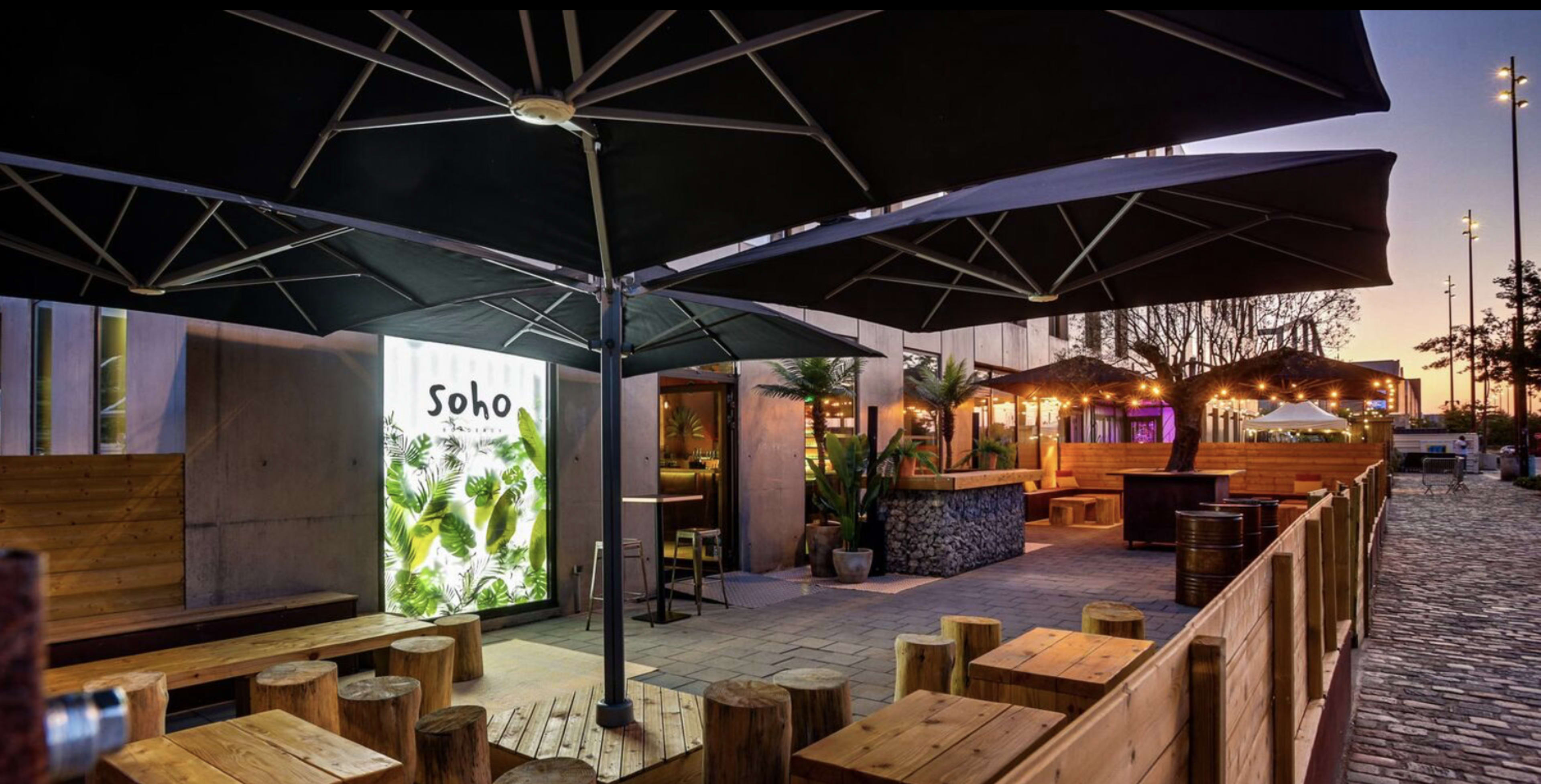 The image shows an outdoor seating area of a venue called "Soho," featuring wooden benches, large umbrellas, and lush greenery, illuminated by warm lights during twilight.