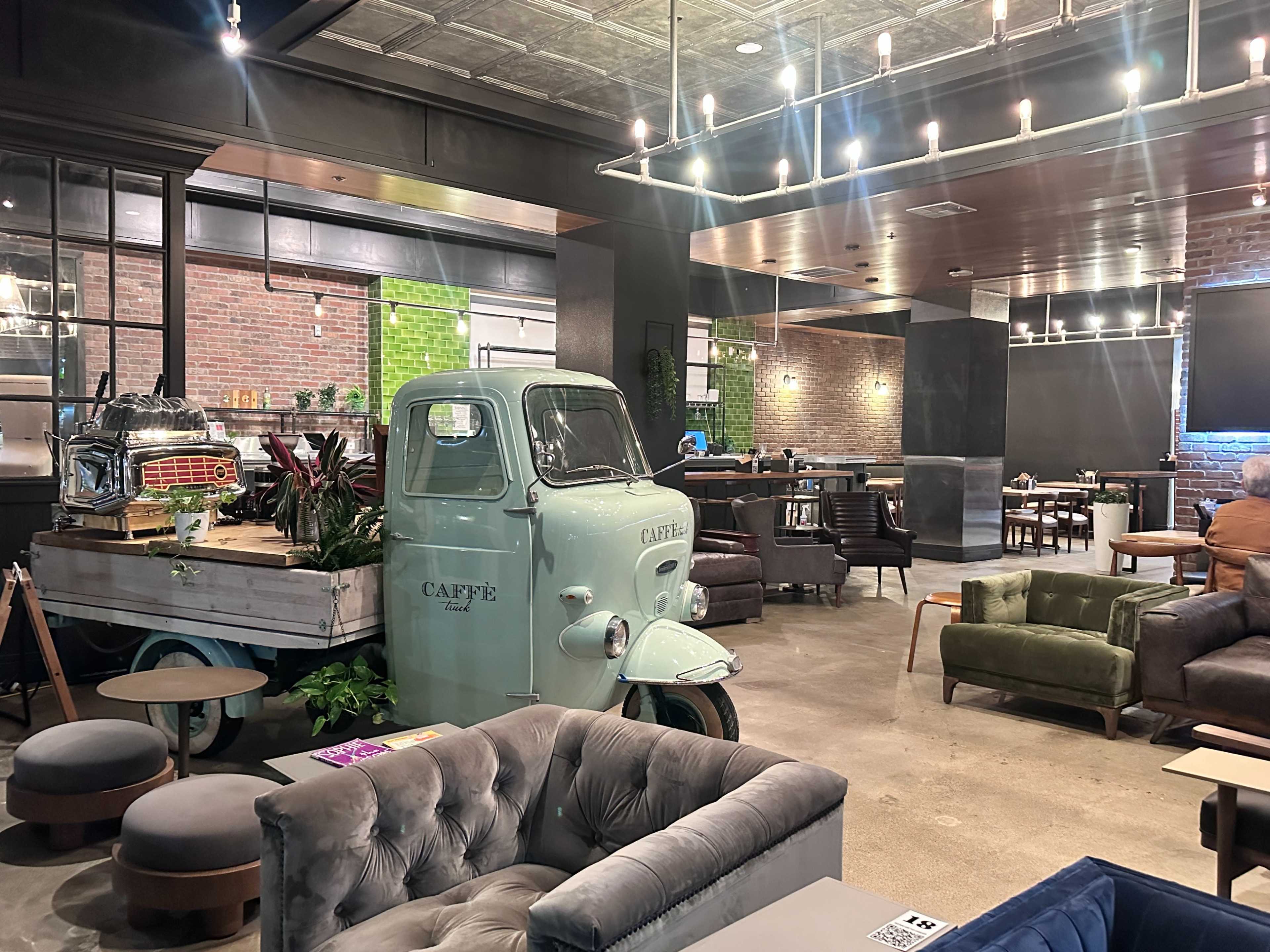 The image shows an indoor café space featuring a vintage truck converted into a display area, surrounded by various seating options and modern decor.