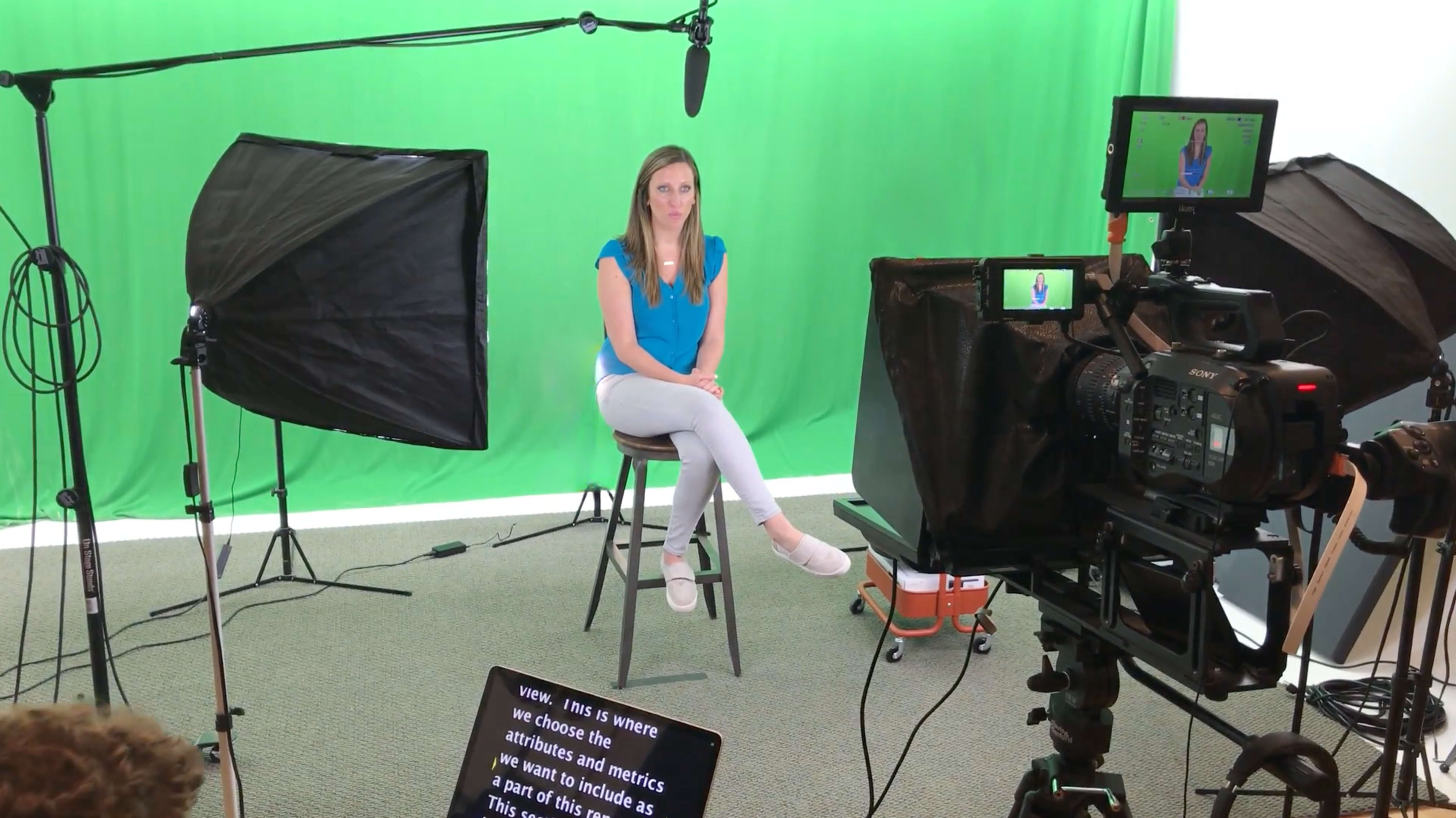 A woman sits on a stool in front of a green screen, surrounded by lighting equipment and a camera setup.