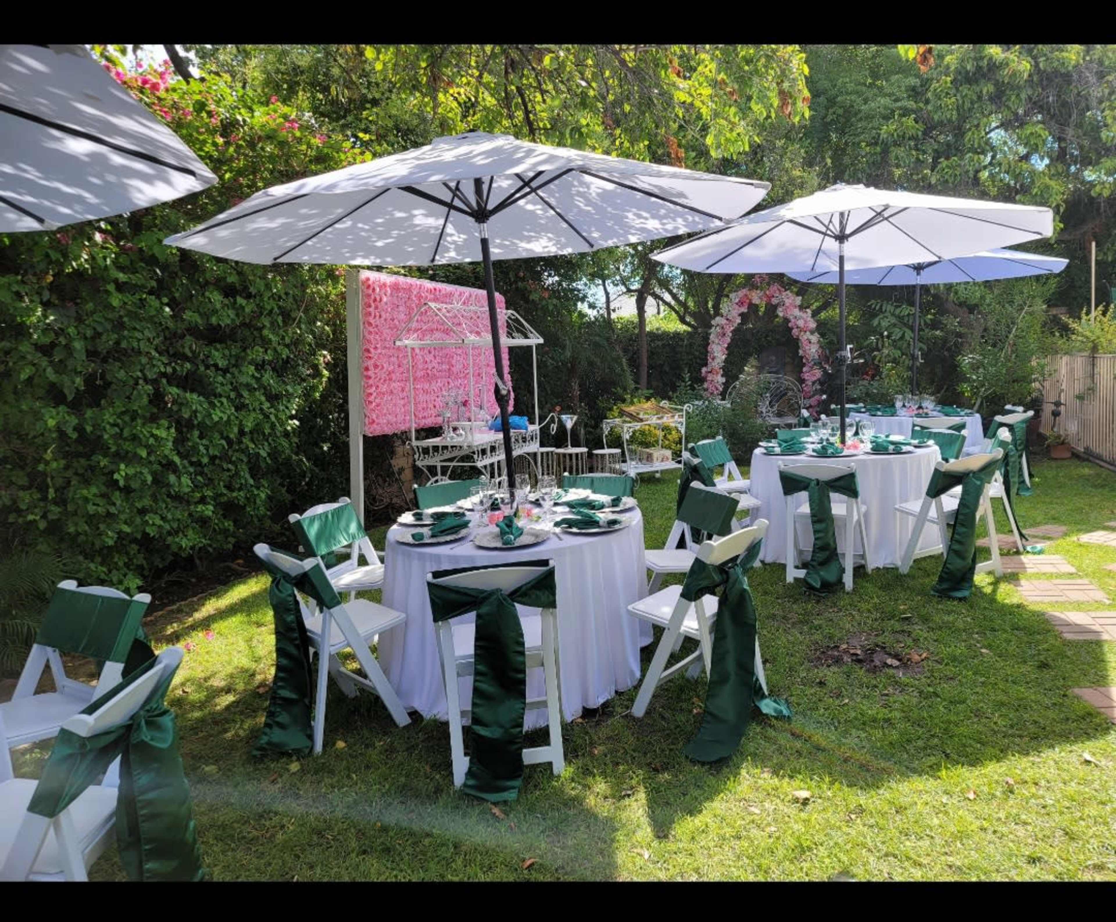 A garden setting for an event, featuring several white tables with green sashes, large umbrellas for shade, and decorative elements in the background.
