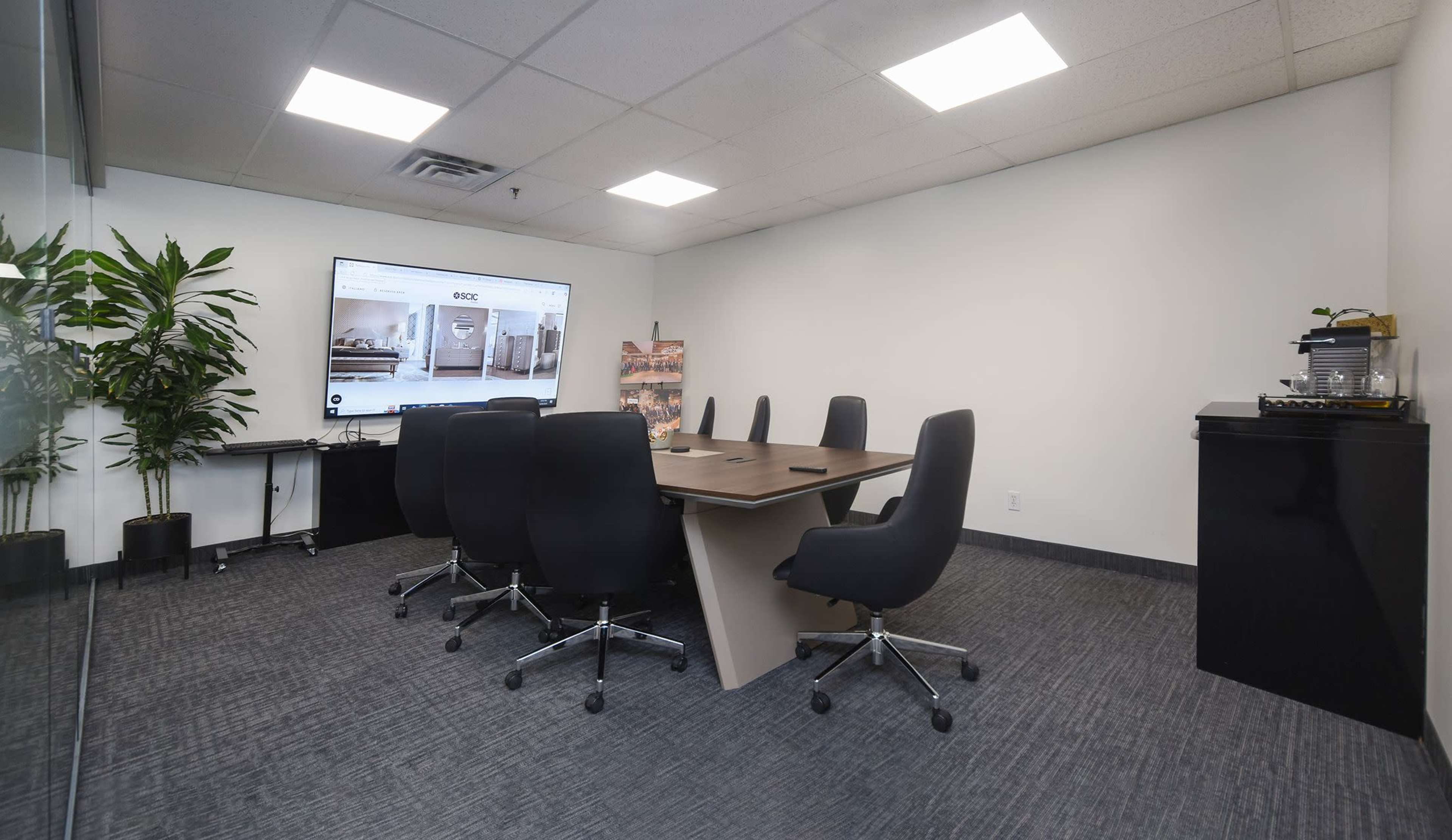 A modern conference room features a large table surrounded by six black office chairs, with a wall-mounted screen displaying a webpage and a potted plant in the corner.