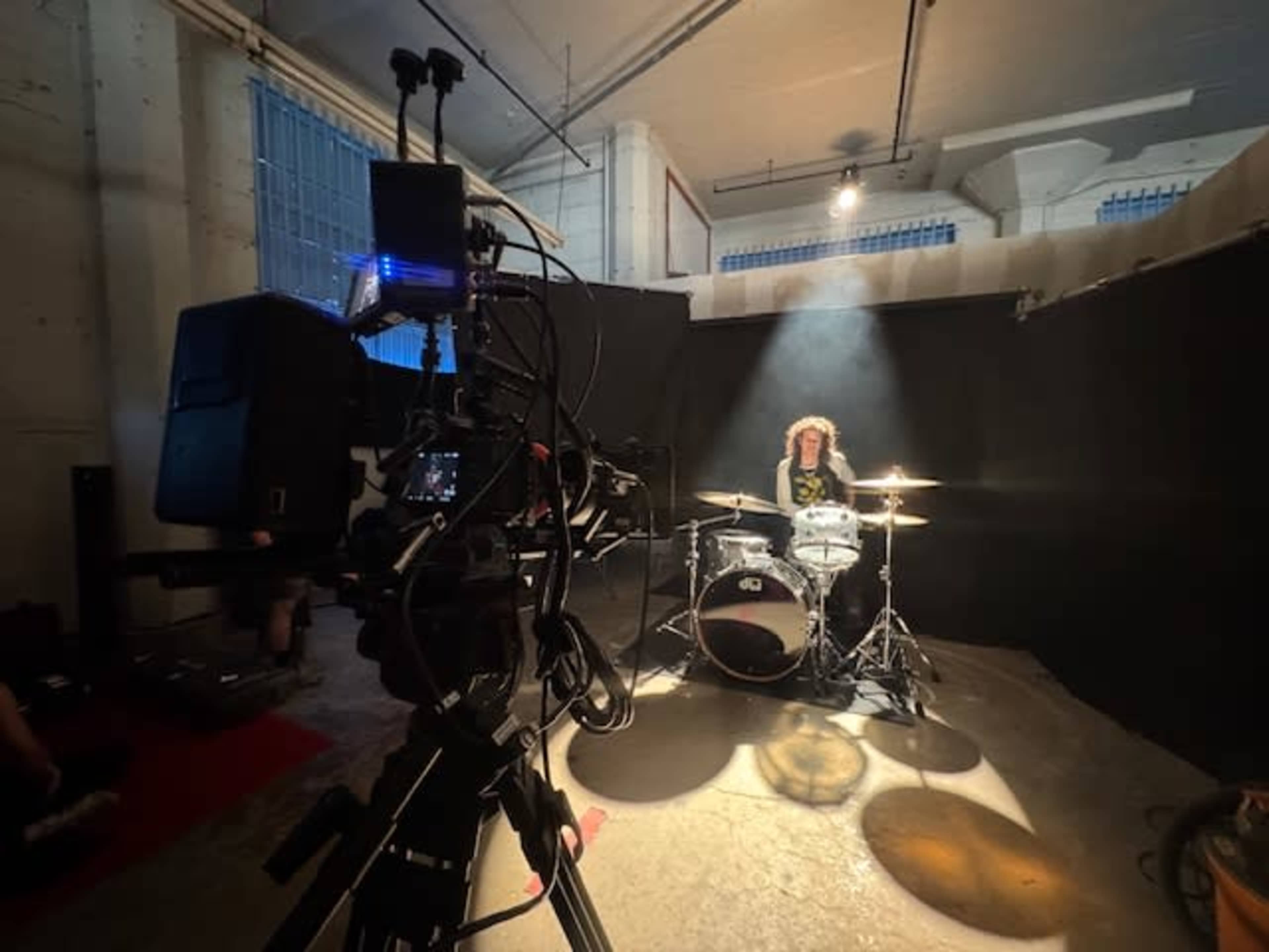 A drummer sits behind a drum set in a dimly lit room, with spotlighting illuminating the drummer against a backdrop of black curtains.
