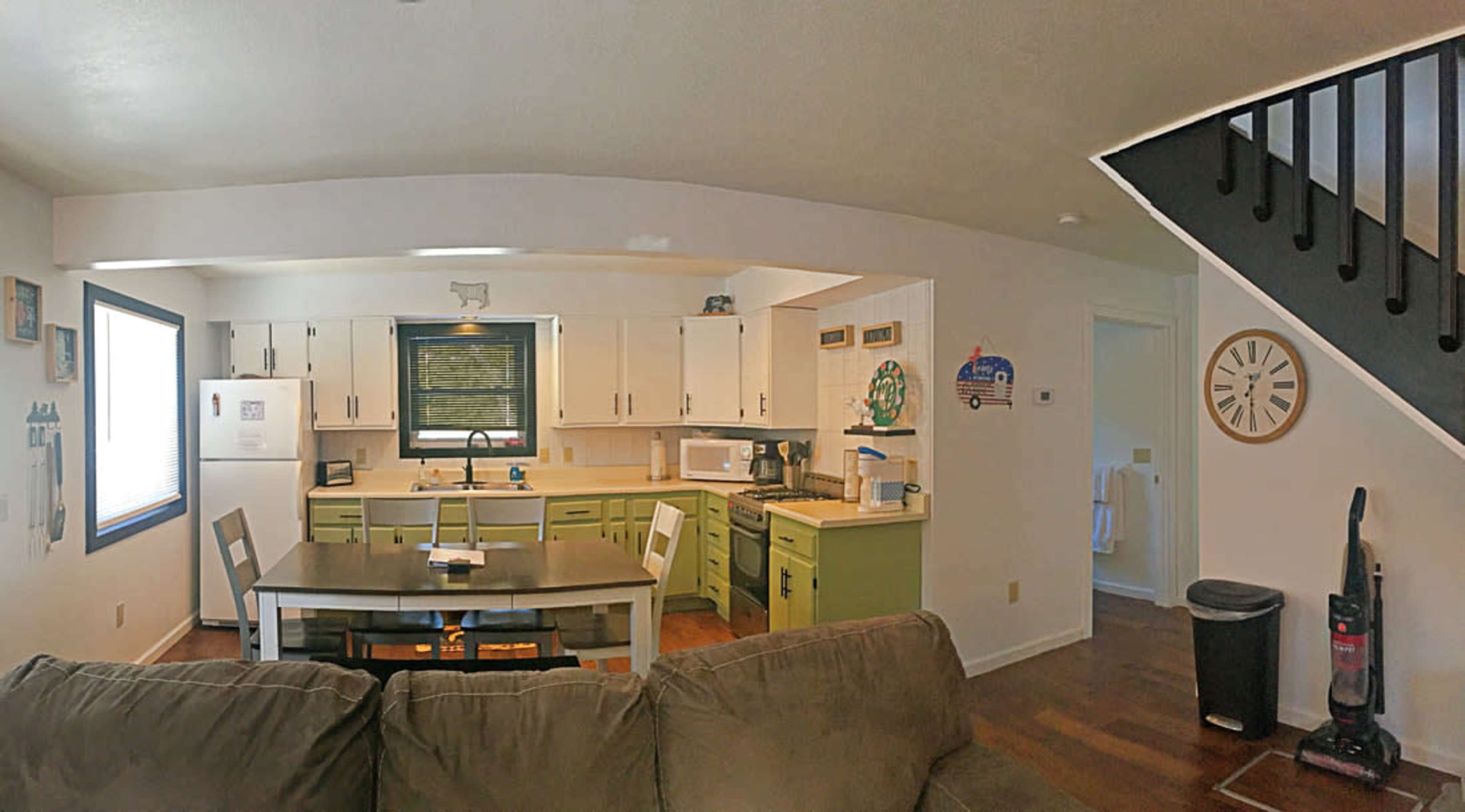 A bright, open-concept kitchen and dining area with green cabinetry, a white refrigerator, and a staircase in the background.