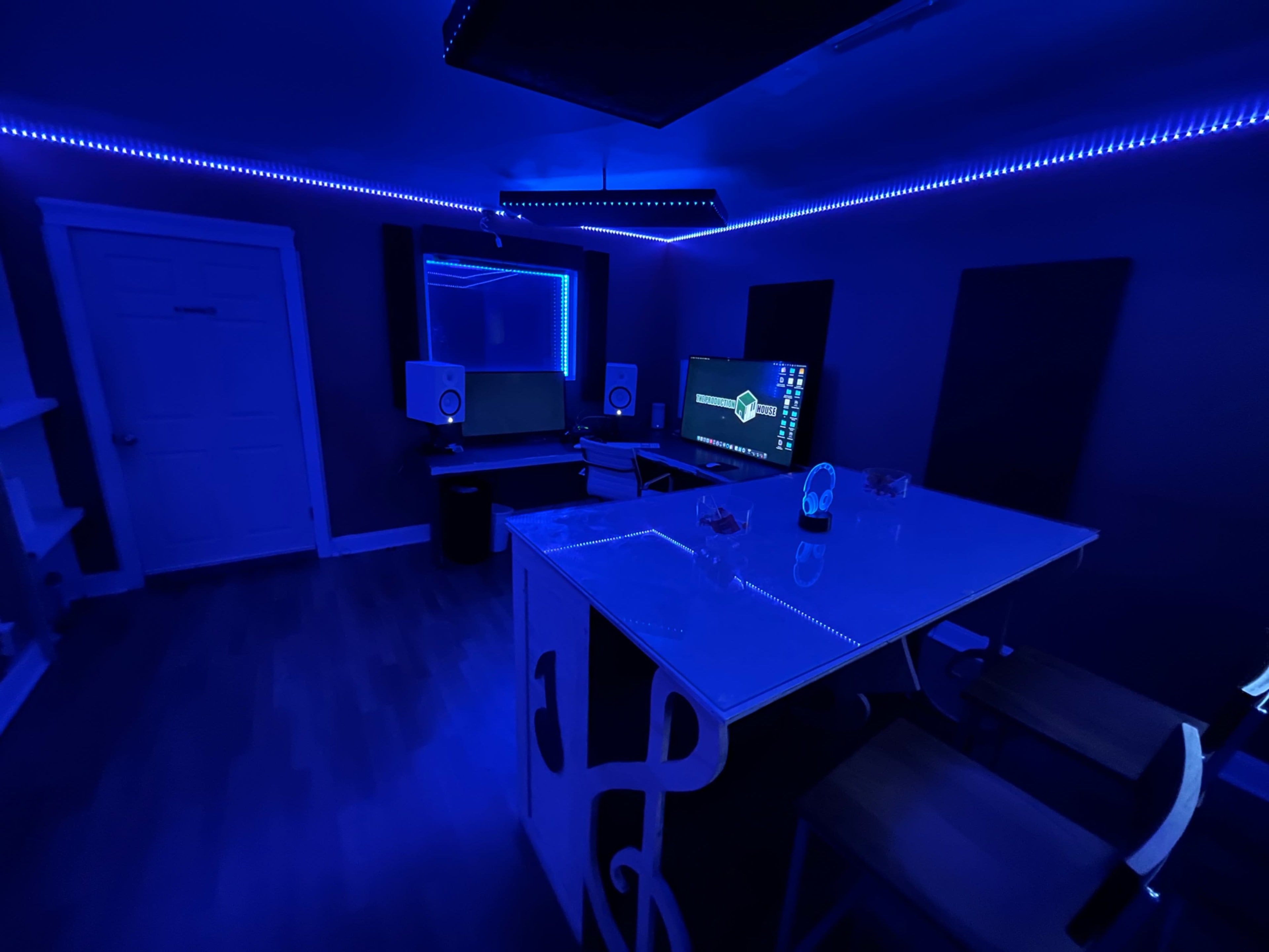 A dimly lit room with blue LED lights, featuring a desk with monitors, speakers, and a large table.