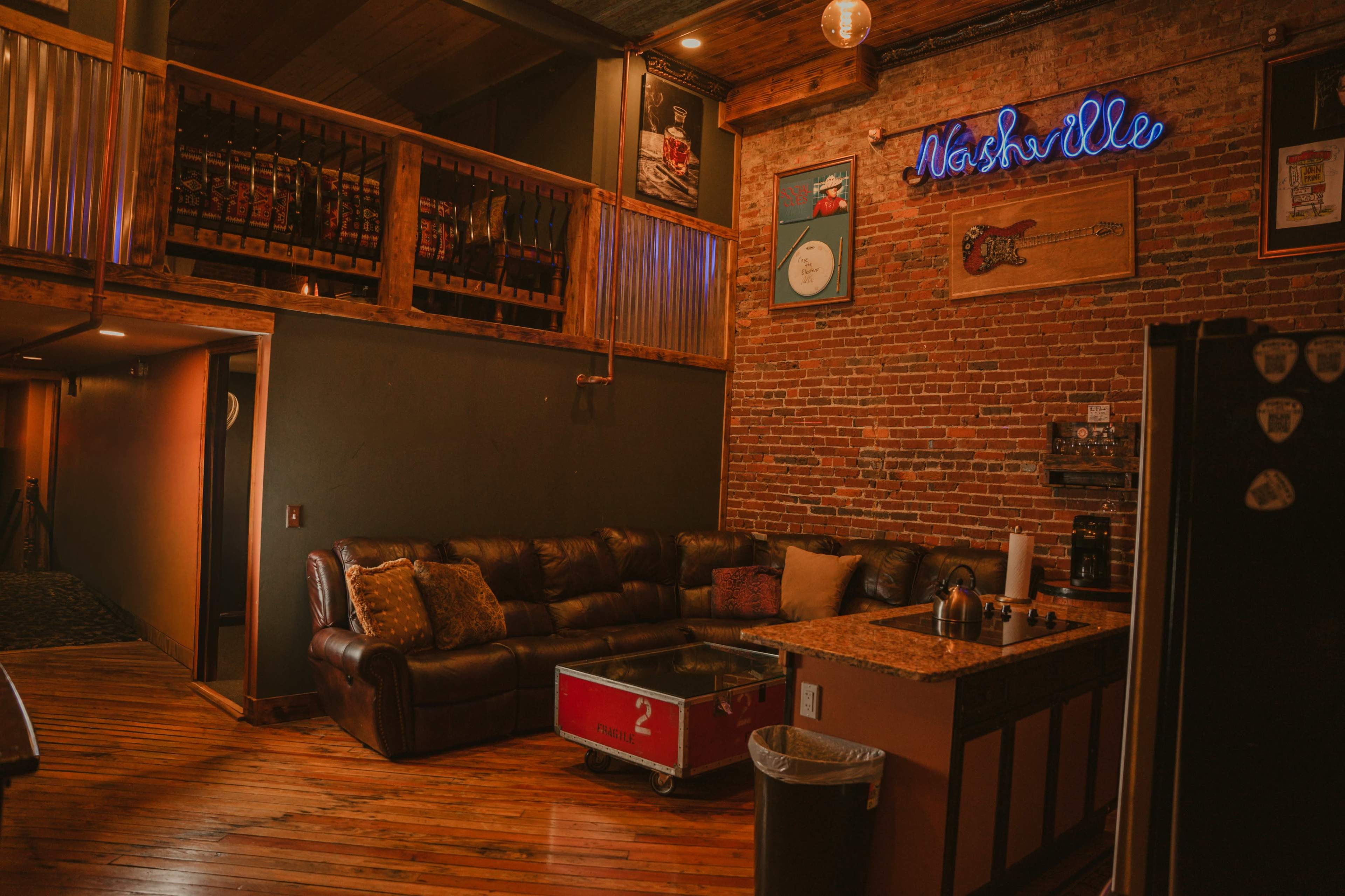 The image shows a rustic interior with exposed brick walls, a leather sofa, and a wooden coffee table in a cozy lounge area, featuring a neon "Nashville" sign and a well-stocked shelf above.