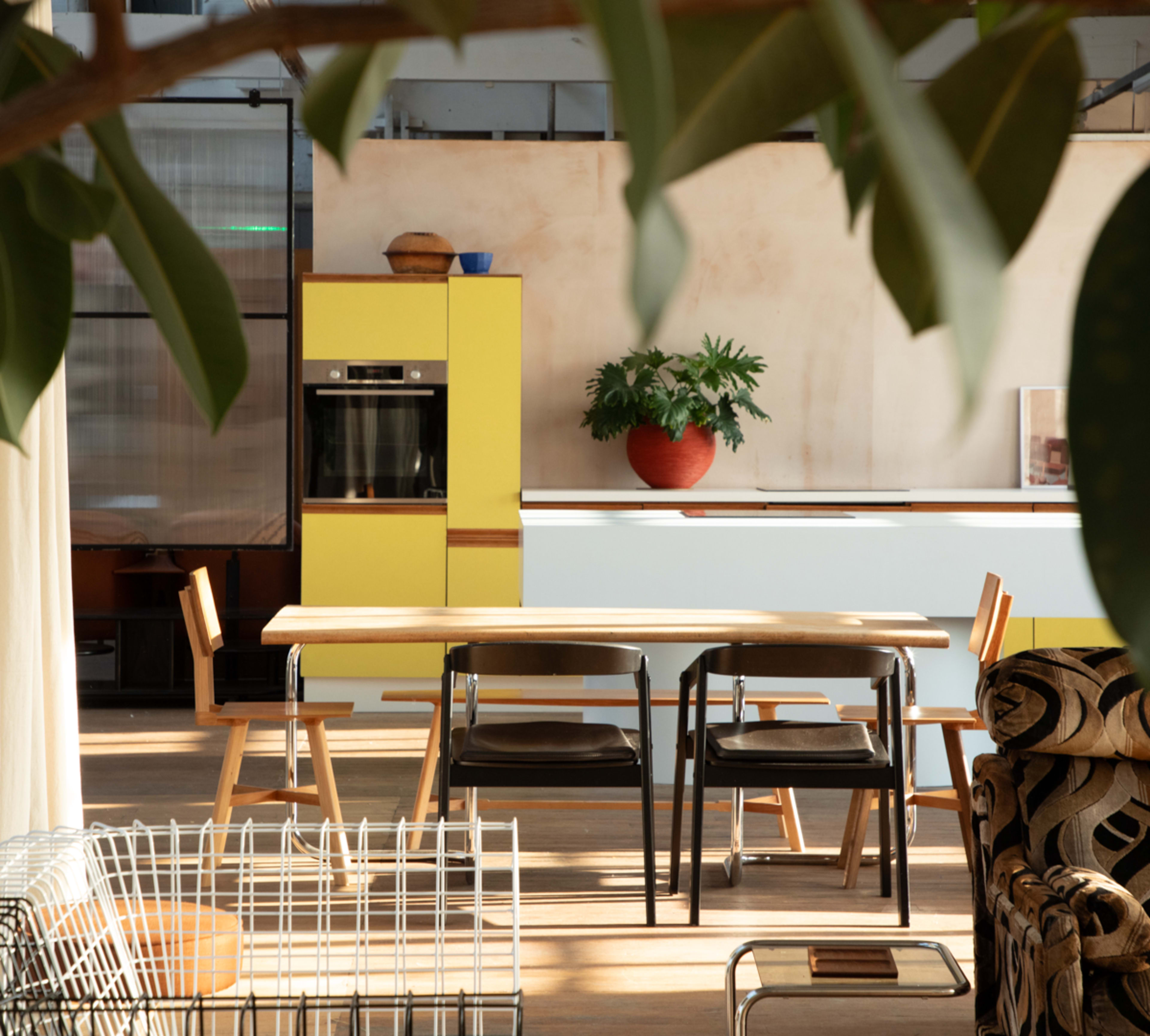A modern kitchen with a yellow cabinet, a wooden dining table, and several chairs surrounded by greenery.