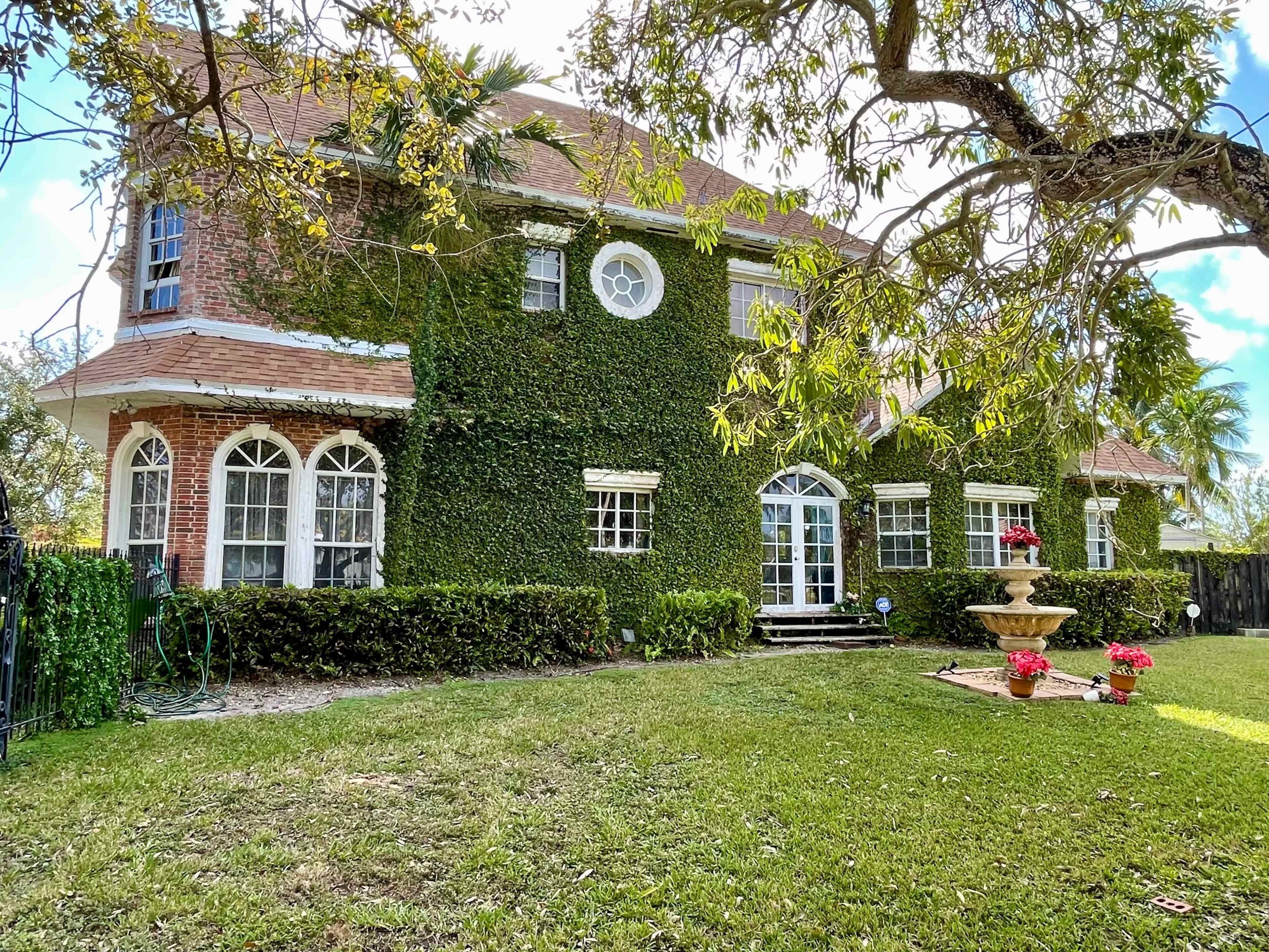 A two-story brick house covered in ivy is surrounded by a well-maintained lawn and features a central fountain with potted flowers in the front yard.