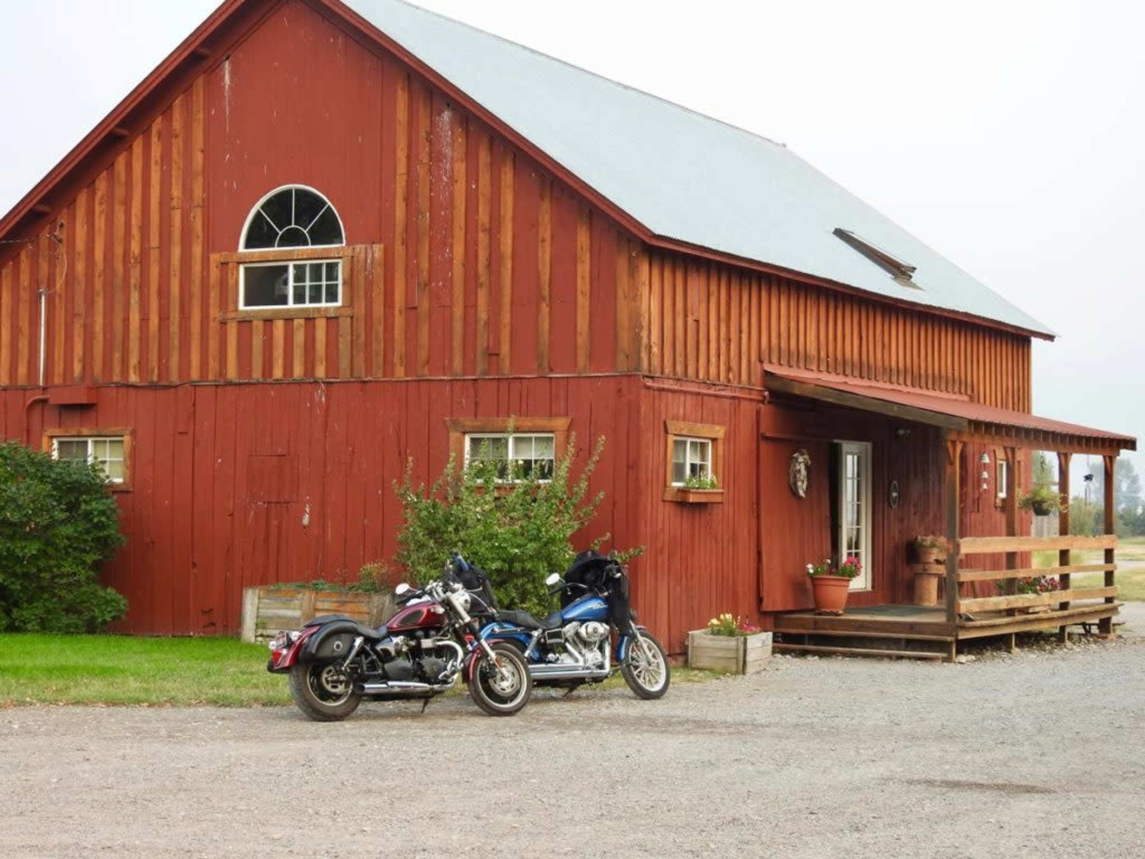 Two motorcycles are parked in front of a red barn with a porch and several windows.