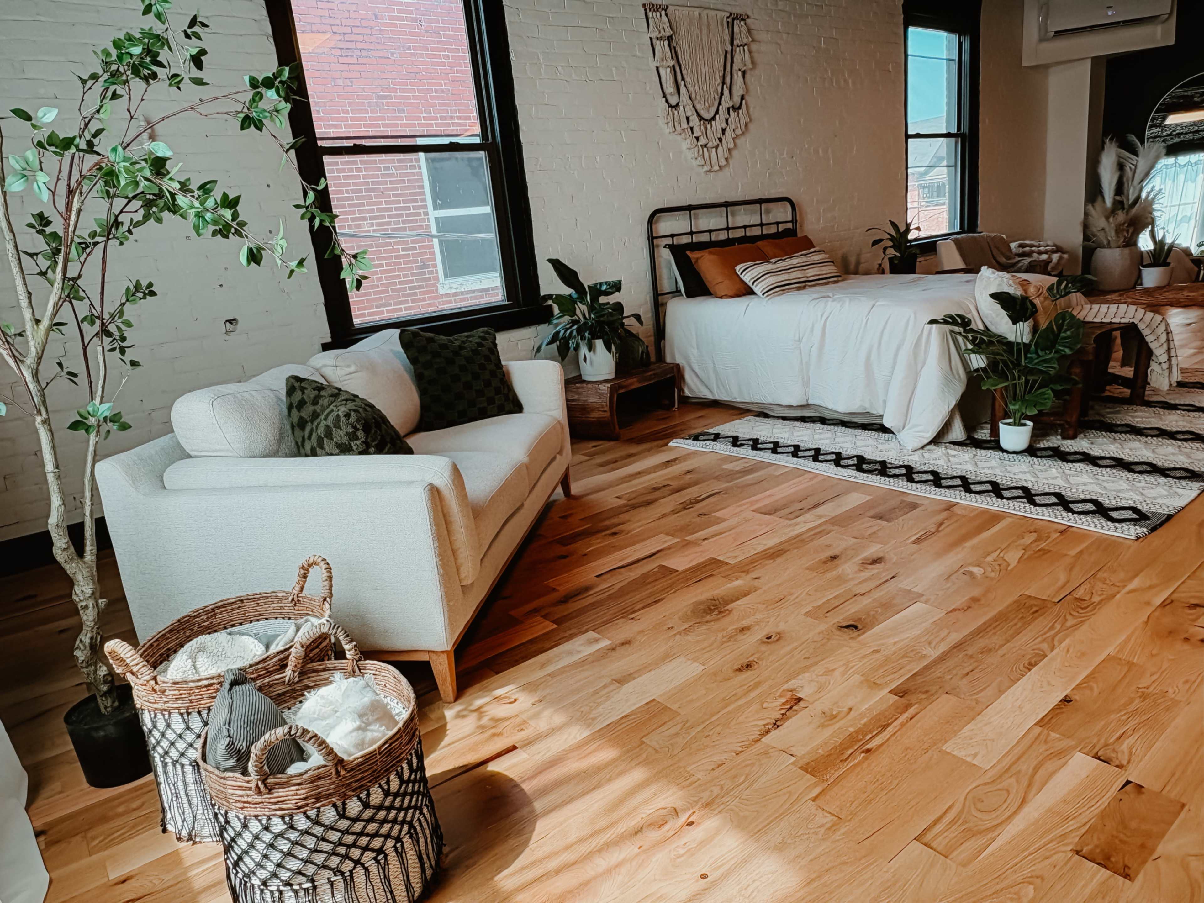 The image shows a spacious bedroom featuring a bed, a cozy seating area with a light-colored sofa, plants, and hardwood flooring.