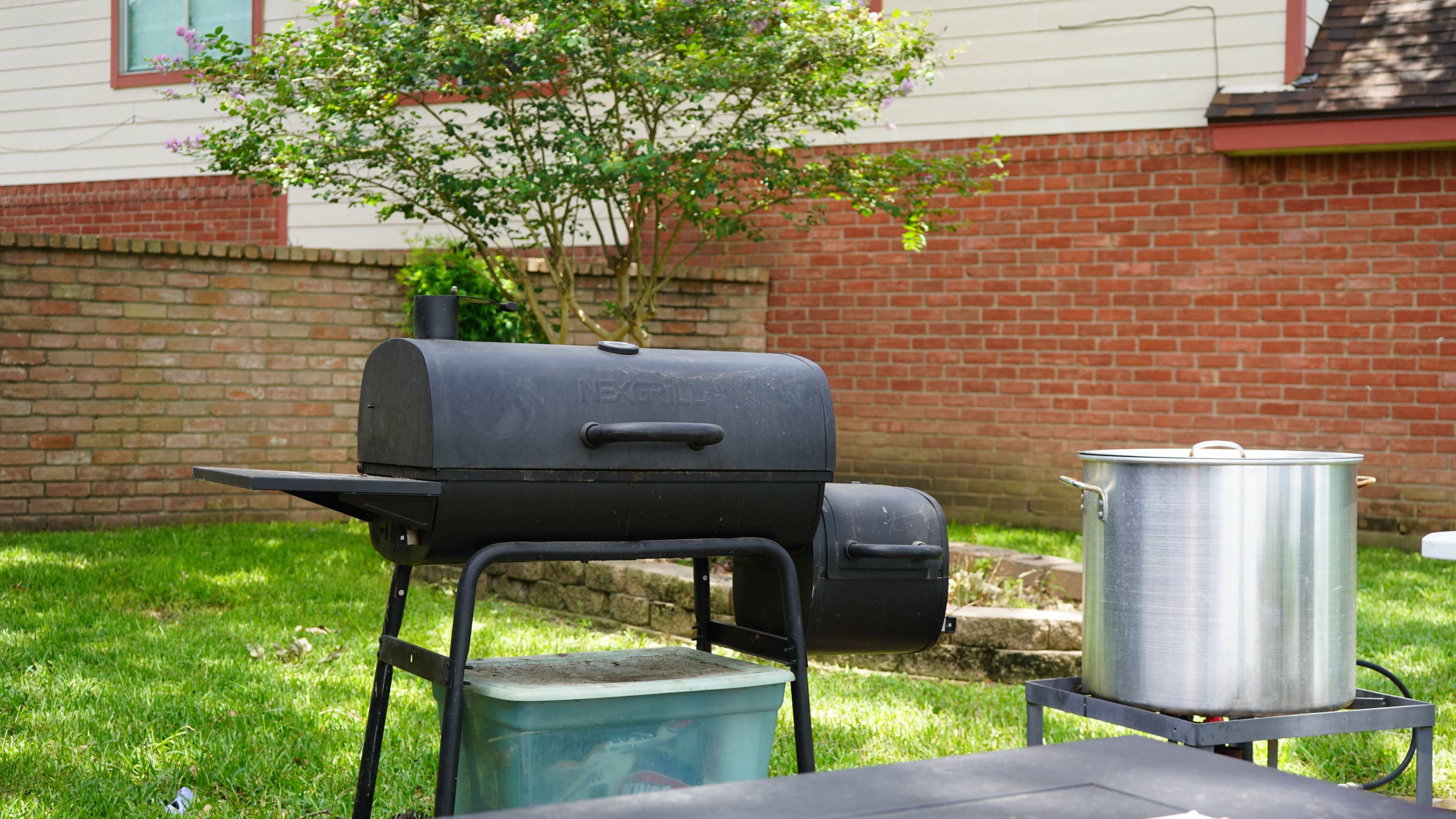 A black grill sits on a stand next to a large pot in a grassy backyard with a brick wall in the background.