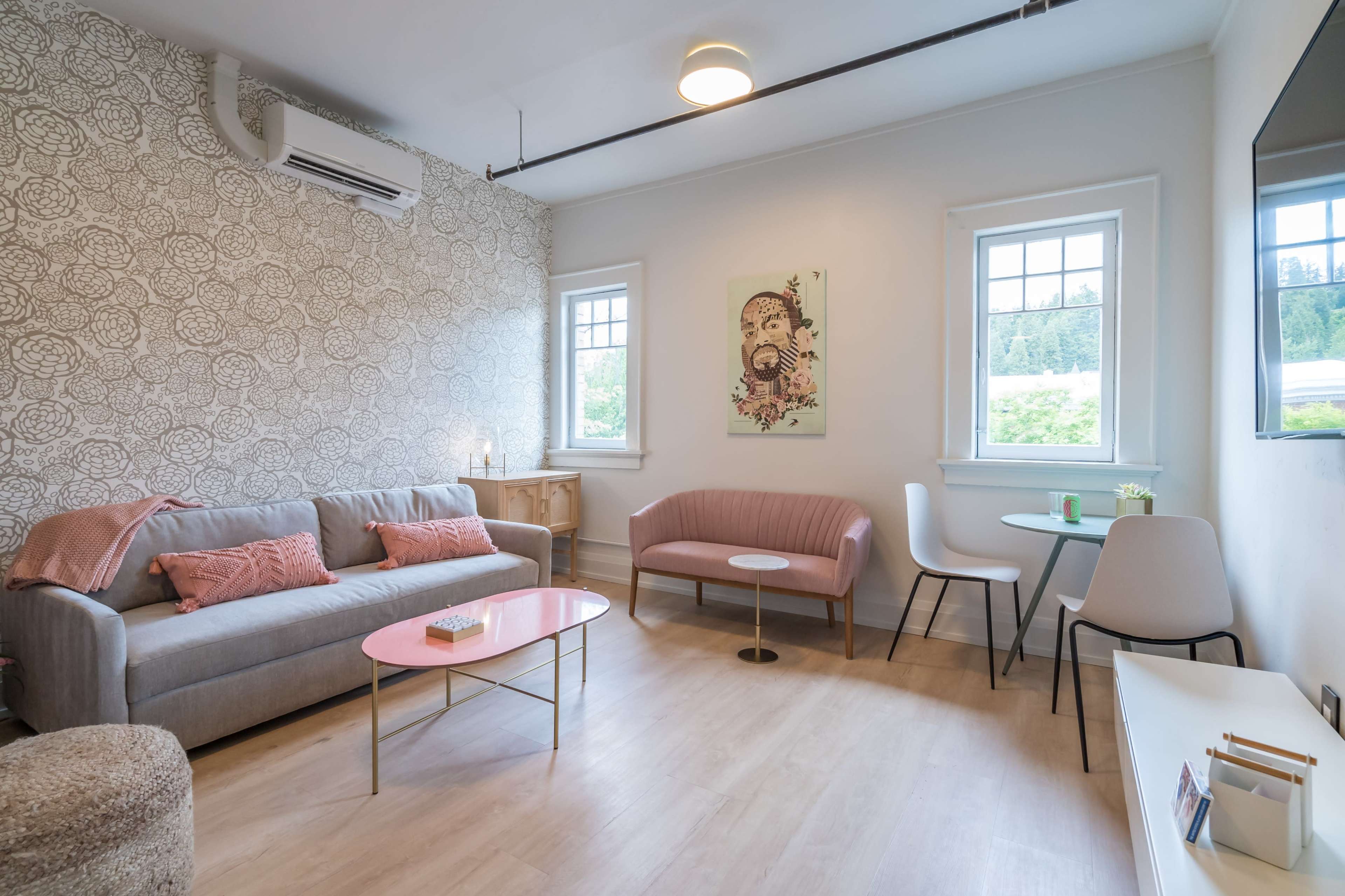 A living room with a gray sofa, a pink chair, a light pink coffee table, and two windows overlooking greenery.