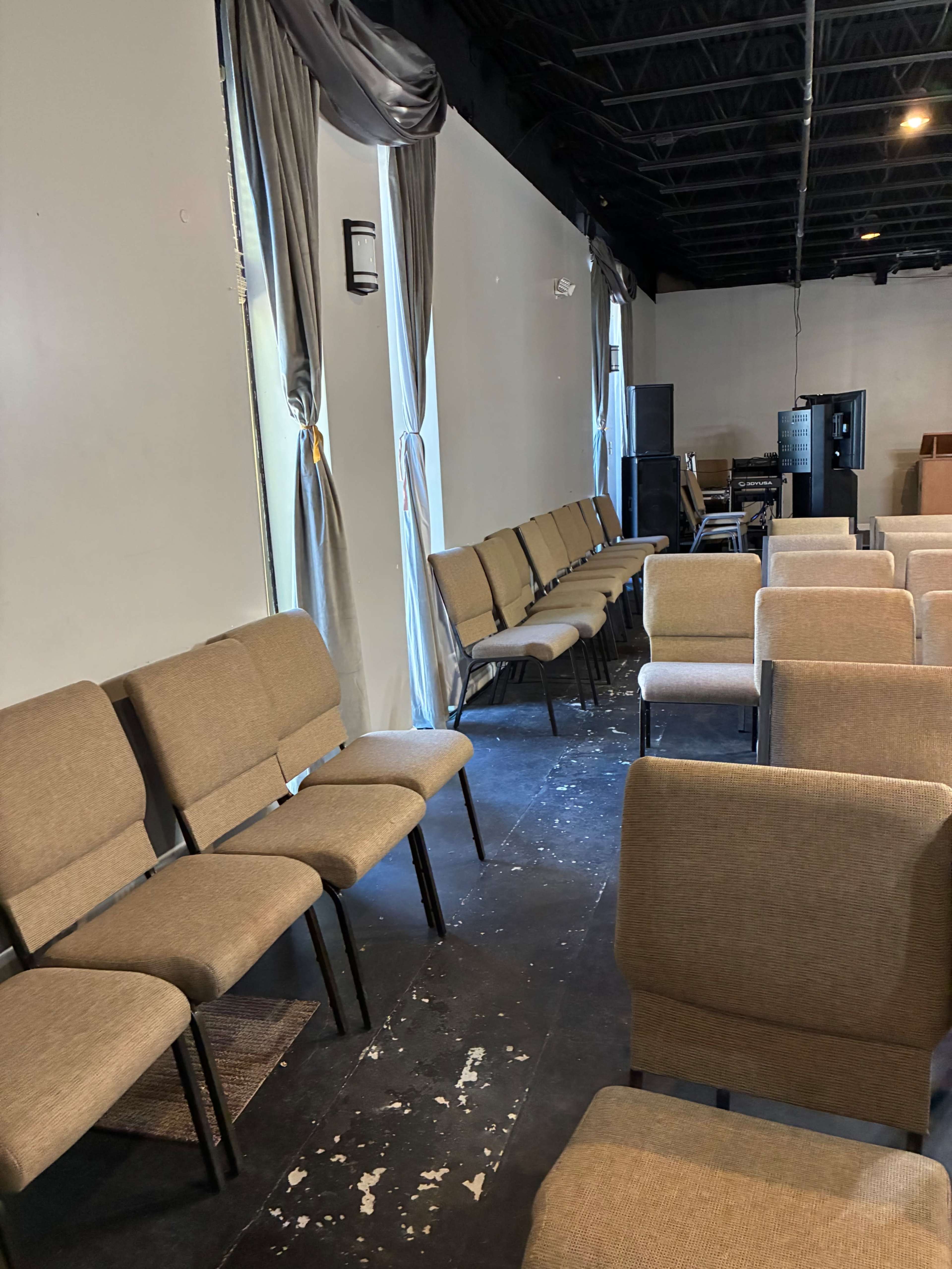 The image shows a partially filled room with rows of chairs arranged neatly in front of a wall, with some curtains hanging by the windows.