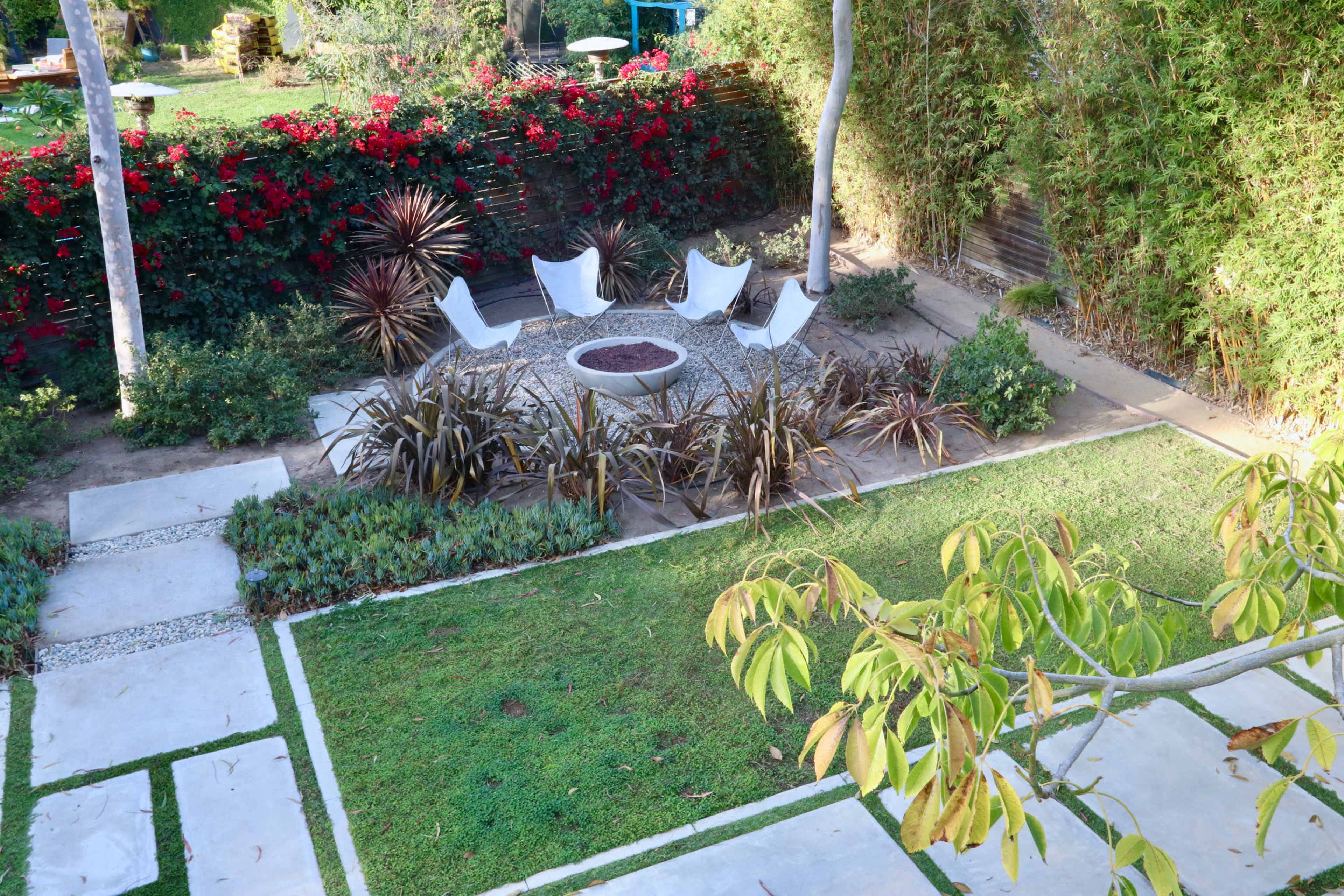 A landscaped garden featuring a circular seating arrangement with four chairs around a central fire pit, surrounded by a variety of plants and pathways.