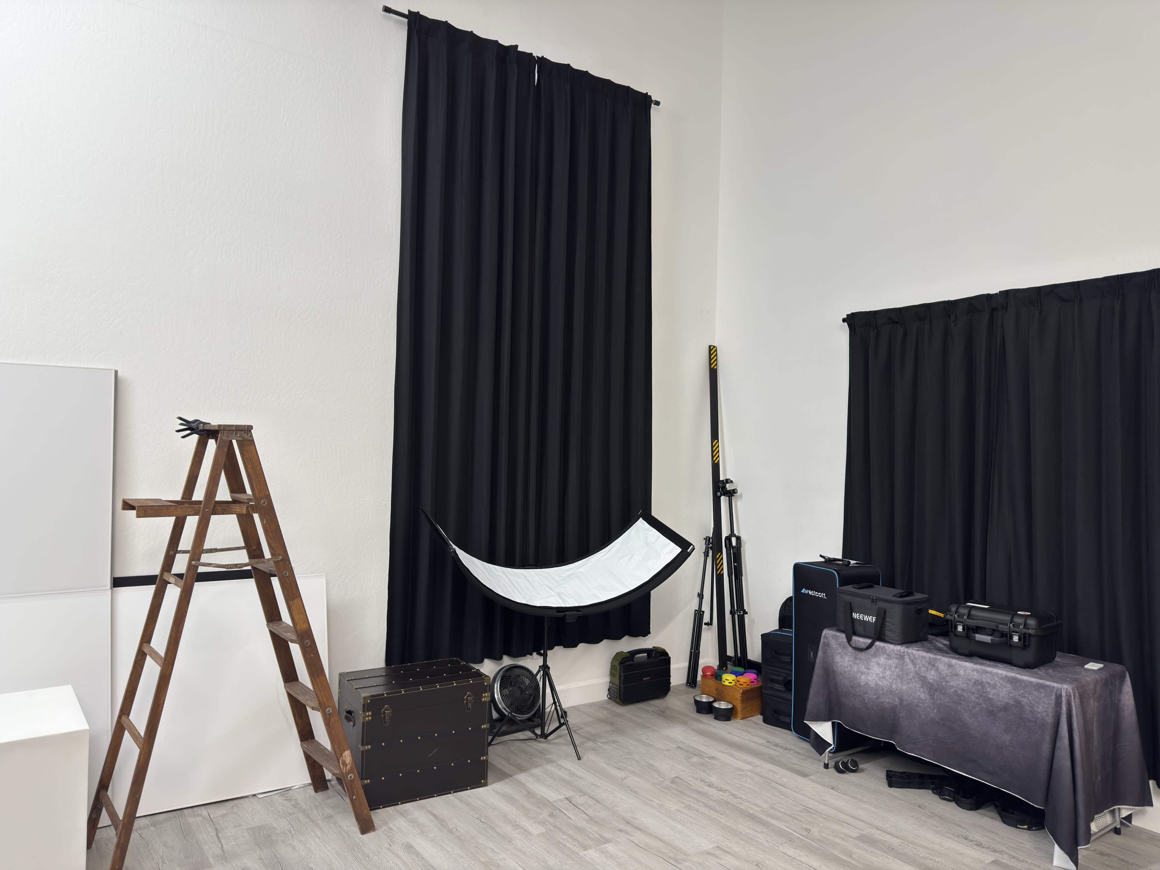 The image shows a studio space with a wooden ladder, a black curtain, a white reflector, and various equipment, including a trunk and speaker boxes.